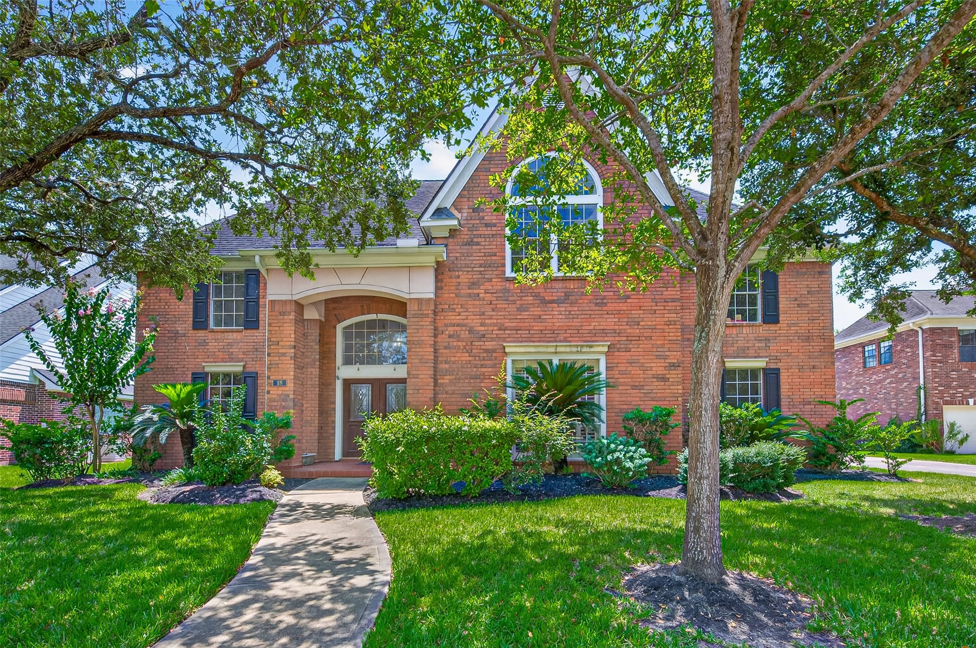 18 Tredington Street, Sugar Land, TX 77479 Elegant two-story brick home with charming arched entryway, surrounded by lush landscaping and shaded by mature trees.