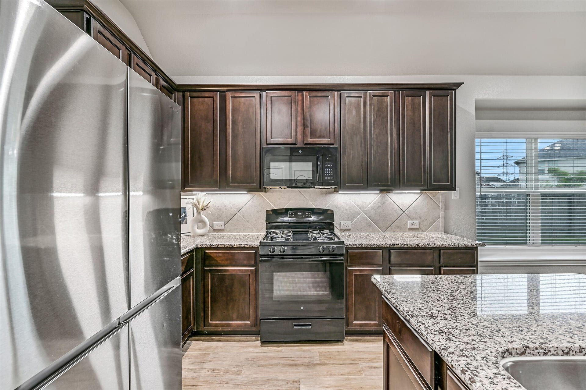 Granite countertops continue throughout the workspace, complemented by a gas range, built-in microwave, tile backsplash, under-cabinet lighting, and stainless refrigerator. Everything is thoughtfully arranged for both functionality and style.
