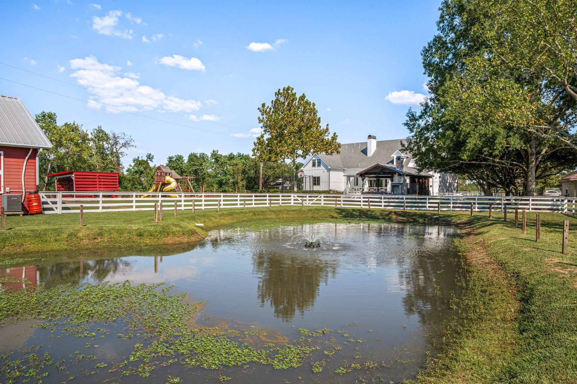 6571 Manorwood Drive, Katy, TX 77493 This serene, stockable pond with bluegill, catfish, and largemouth bass features a central aeration fountain and classic white ranch fencing, creating a picturesque centerpiece for the property.