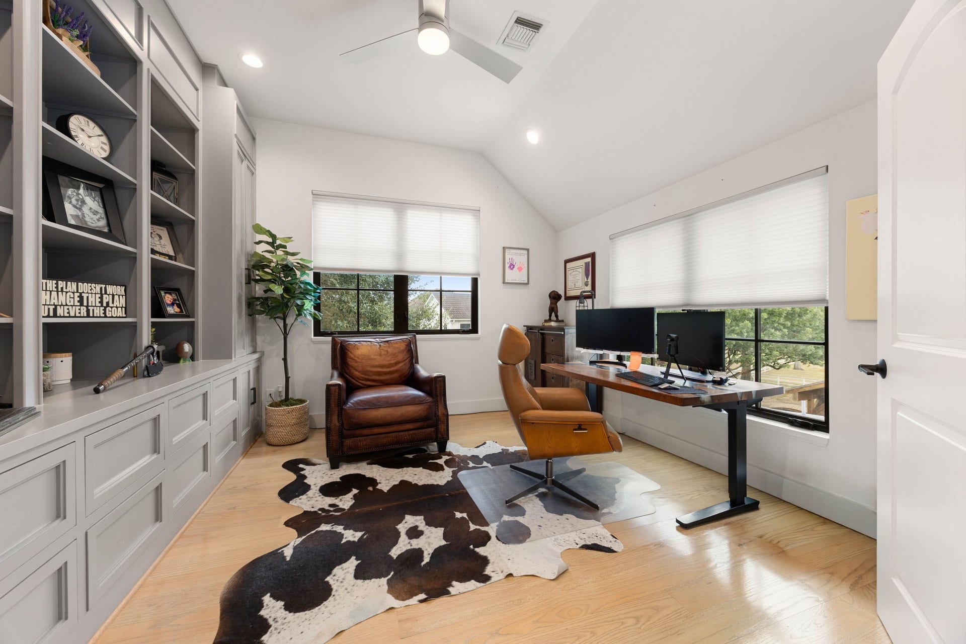 6571 Manorwood Drive, Katy, TX 77493 The office boasts a distinct vaulted ceiling design equipped with recessed lighting and a modern ceiling fan, anchored by beautiful wide-plank wood flooring that carries through the entire upper level. Office features a commanding, floor-to-ceiling built-in shelving and storage unit finished in a sophisticated gray, offering abundant open display space and deep lower storage cabinetry.