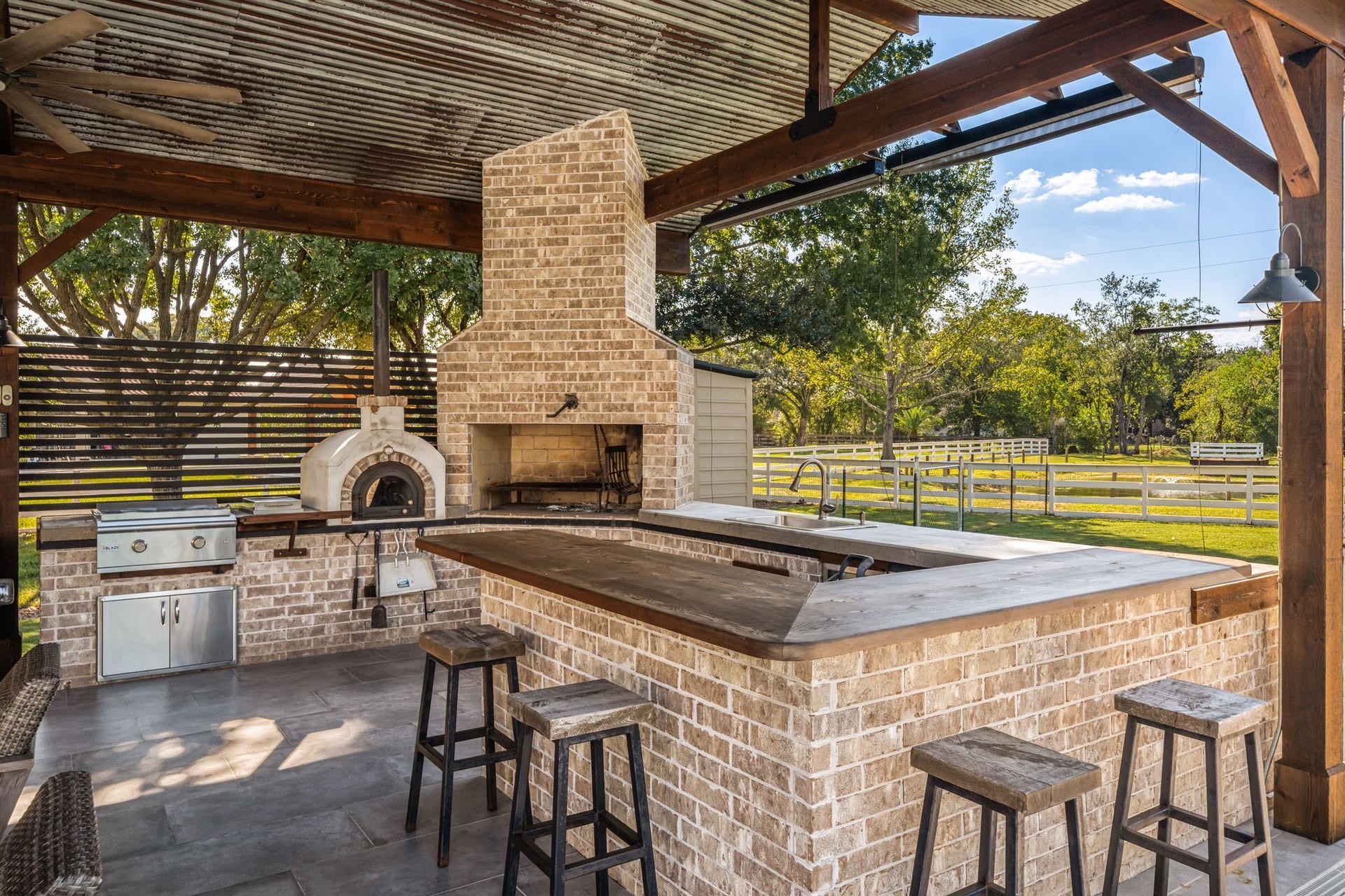 6571 Manorwood Drive, Katy, TX 77493 The multi-tiered brick island offers an expansive wood-topped breakfast bar with seating for guests, a built-in undermount sink, and durable concrete floorsall sheltered beneath a rustic corrugated metal pavilion ceiling with integrated ceiling fans and lighting.