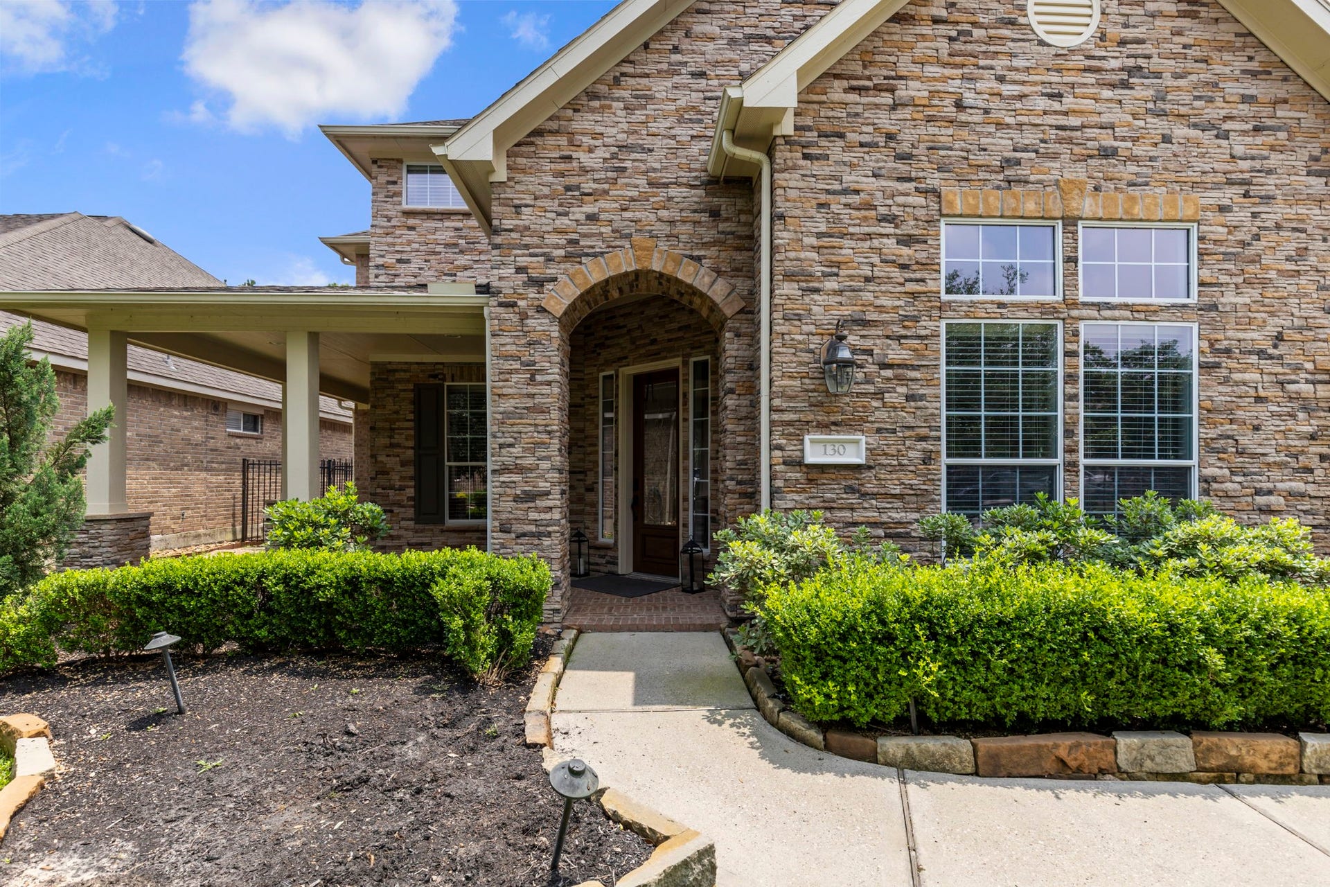 130 S Arrow Canyon Circle, The Woodlands, TX 77389 Charming two-story stone house with large windows, a welcoming arched entryway, and a neatly landscaped front garden.