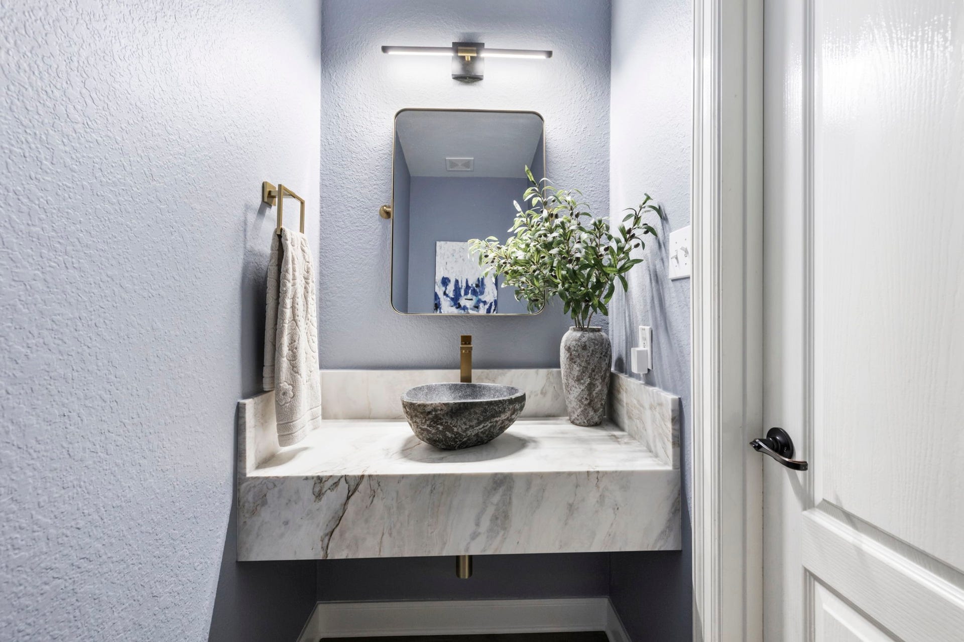 130 S Arrow Canyon Circle, The Woodlands, TX 77389 This is a stylish and modern powder room featuring a sleek stone vessel sink on a marble countertop. It has soft blue walls and elegant lighting.