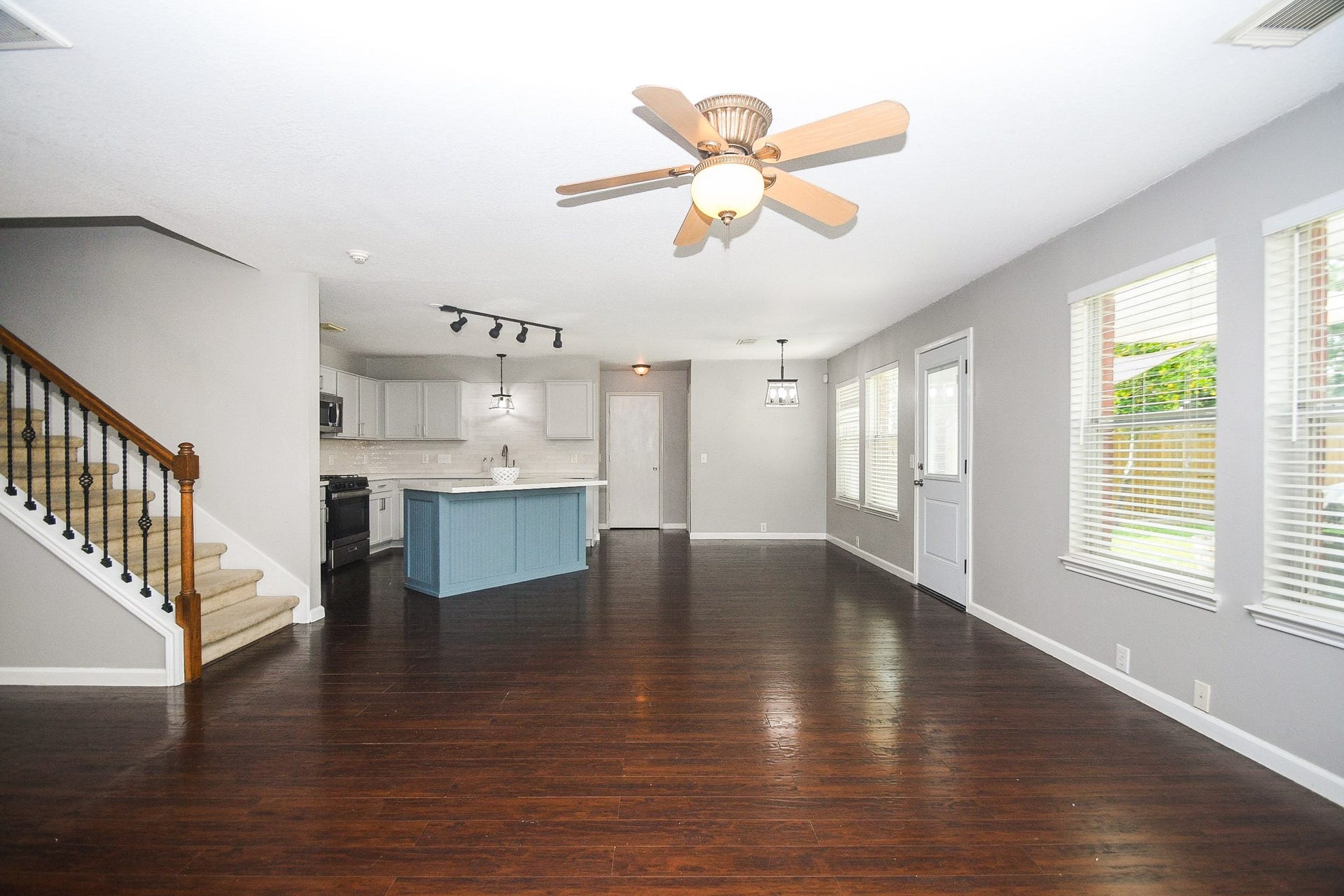 10222 Antelope Alley, Missouri City, TX 77459 Spacious open-concept living area with rich hardwood floors, modern ceiling fan, and abundant natural light from large windows. The kitchen features a stylish blue island, sleek white cabinetry, and stainless steel appliances. Adjacent is a cozy dining area, while a carpeted staircase leads to the upper level.