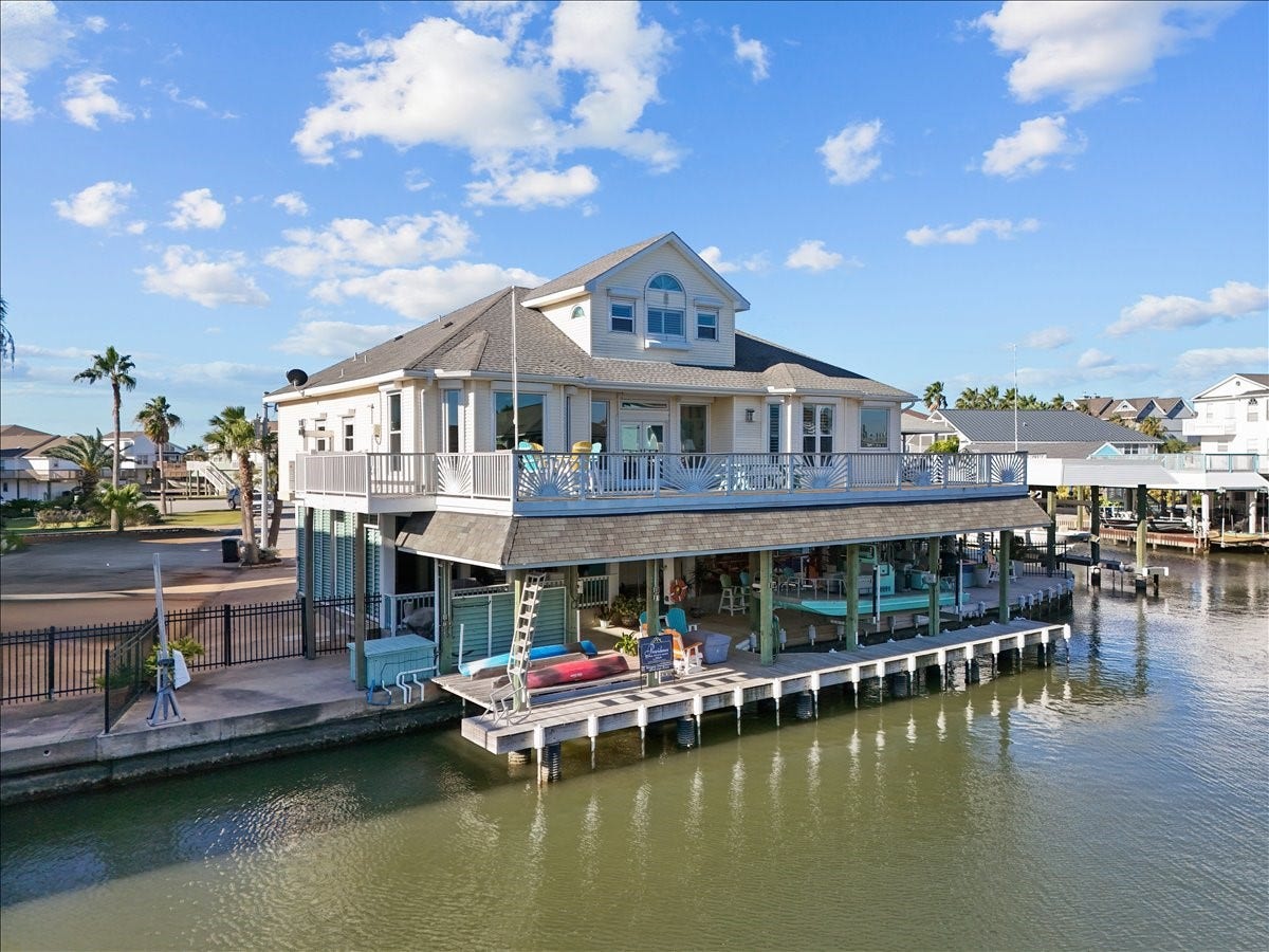 102 Bamaku Bend, Tiki Island, TX 77554 This picturesque view of the back of the home allows you to see the expansive upper deck that has been updated with treated wood, Trex railing and a beautiful sunburst design. There is also a cargo lift on the side of the home that goes down to the ground level next to the garage. The windows have been upgraded and include electric storm shutters except the arched windows.