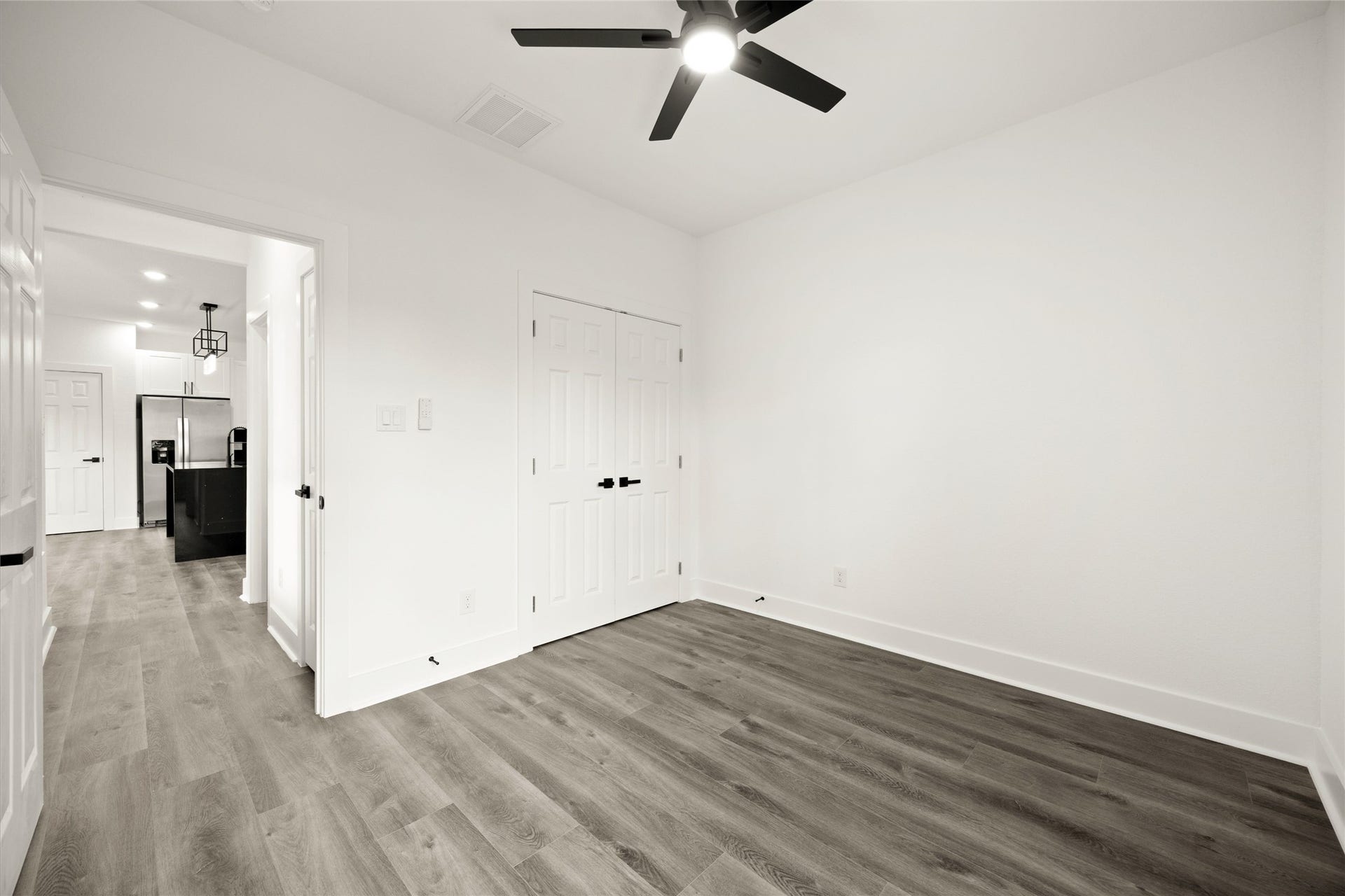 Bright and spacious room with sleek wood flooring, contemporary ceiling fan, and double closet doors, seamlessly flowing into the modern kitchen area.
