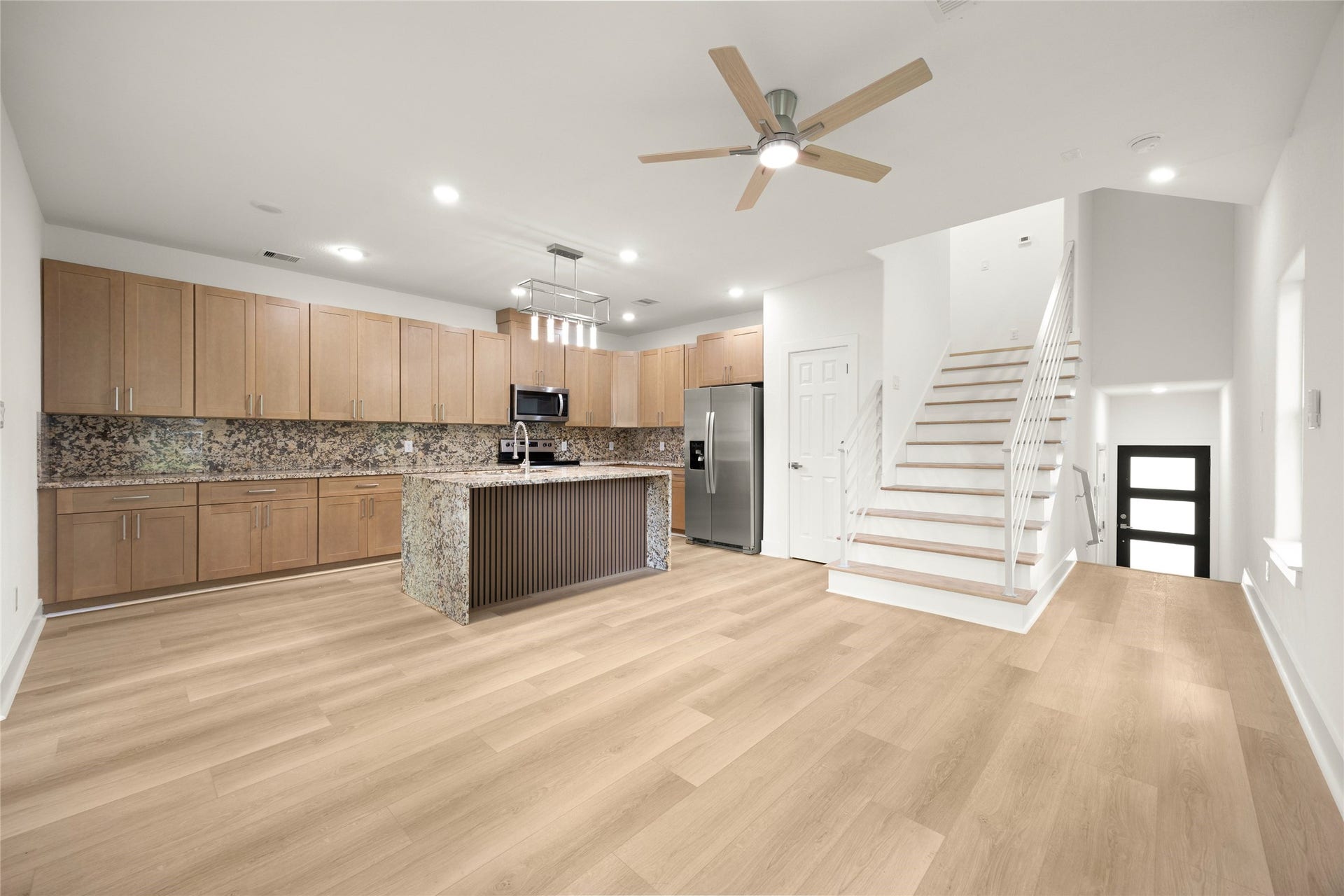 Spacious open-concept kitchen and living area featuring modern wood cabinetry, a large granite island, stainless steel appliances, and sleek wood flooring, complemented by a stylish staircase and ceiling fan.