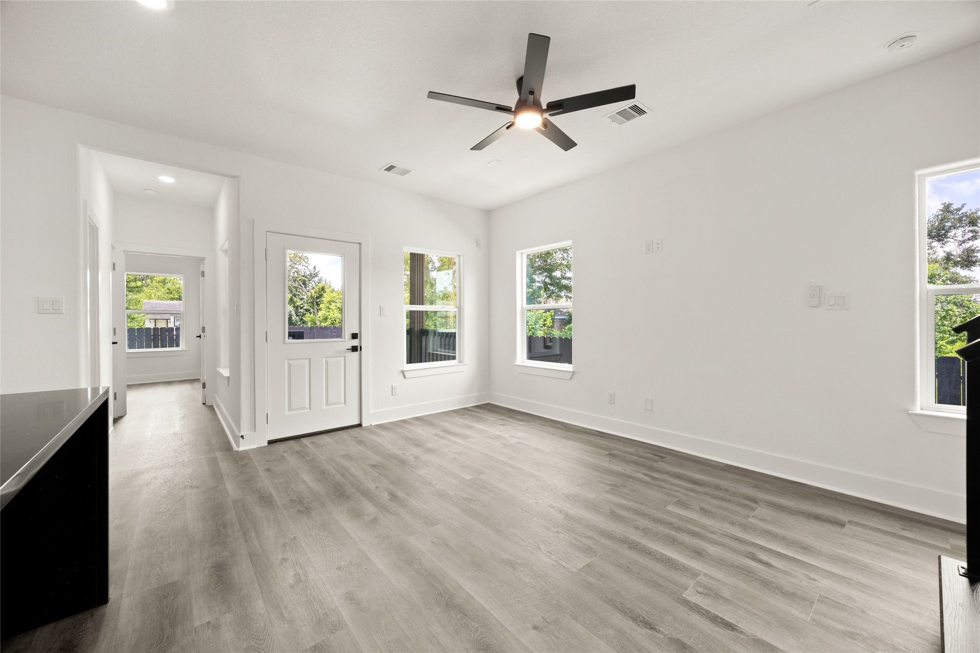 Bright and airy room featuring sleek wood flooring, modern ceiling fan, and ample natural light from large windows, with a view of the lush greenery outside.