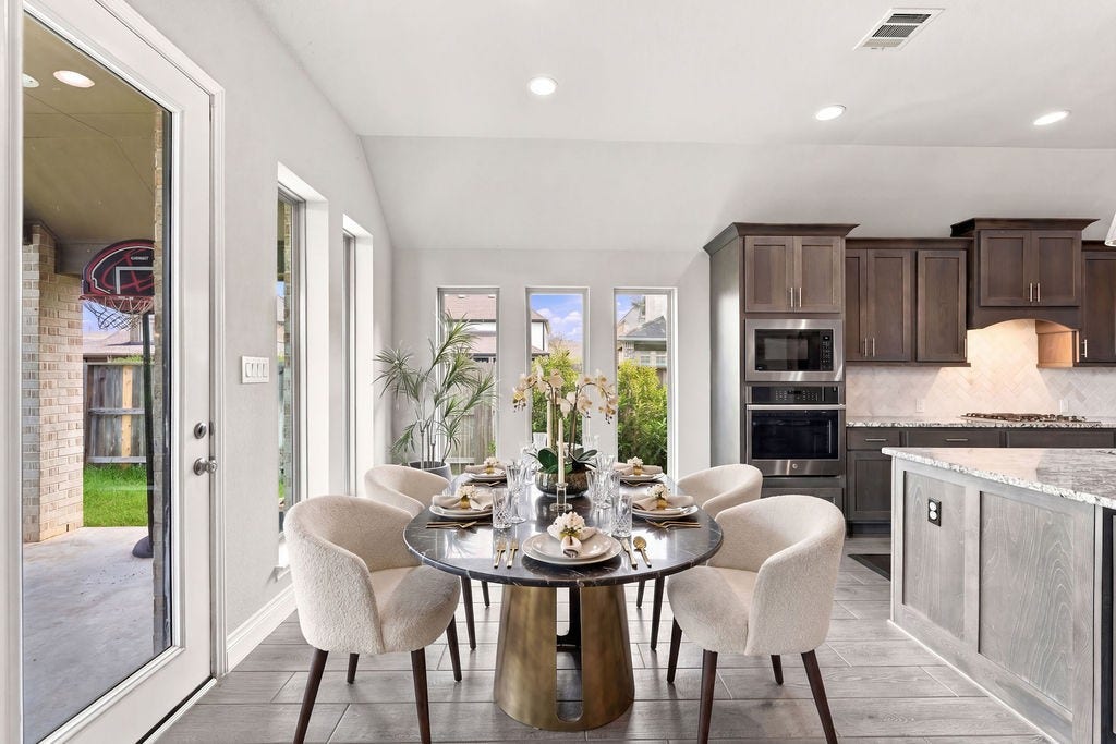 6914 Red Oak Drive, Katy, TX 77493 Bright dining area with a sleek, modern table set for four, seamlessly connected to a stylish kitchen featuring dark wood cabinetry and stainless steel appliances. Large windows and a glass door open to an inviting patio and lush backyard.