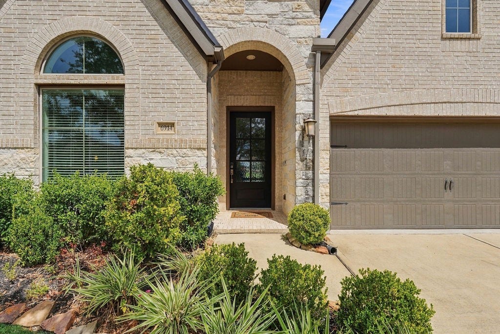 6914 Red Oak Drive, Katy, TX 77493 Charming brick and stone facade with elegant arched entryway, framed by lush landscaping and a spacious garage.