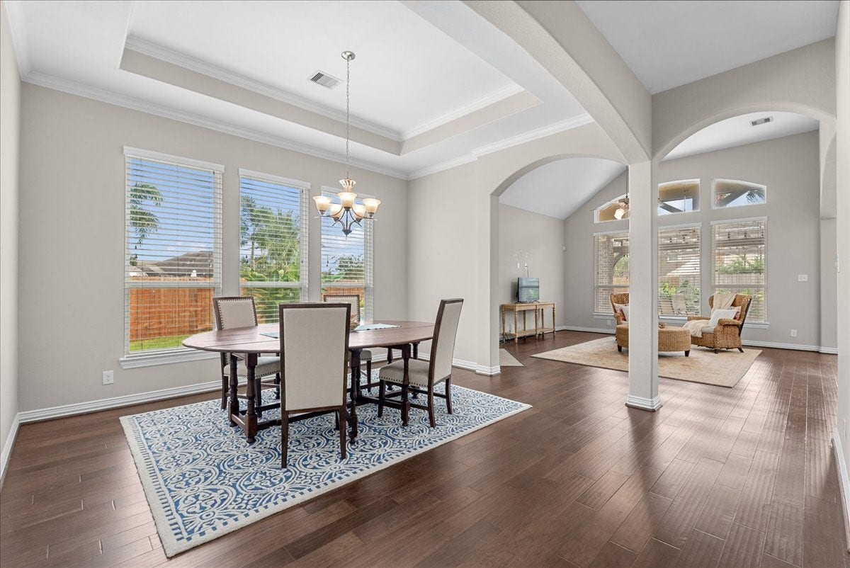 Elegant open-plan dining and living area featuring high ceilings, rich engineered hardwood floors, and abundant natural light from large windows overlooking the backyard.