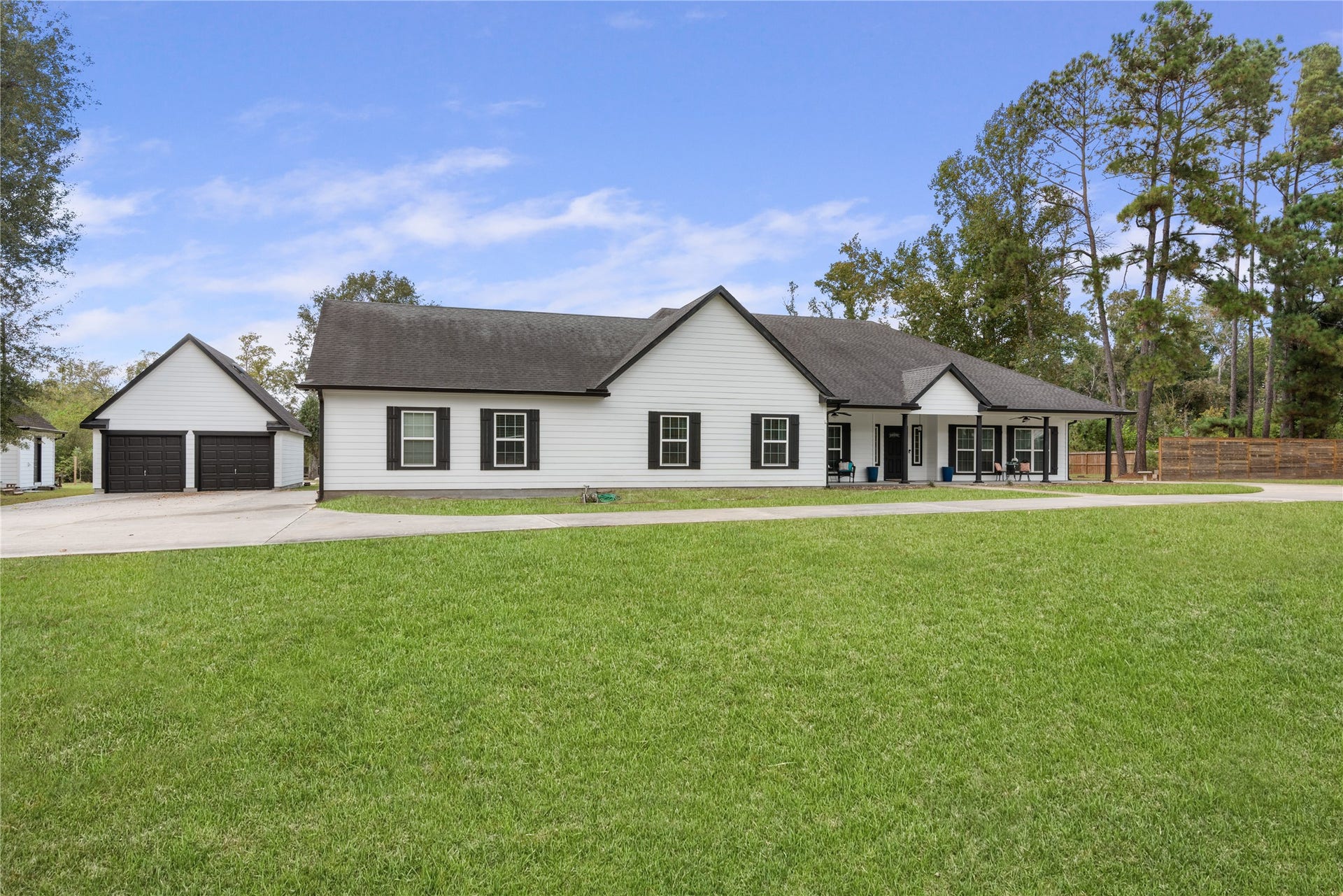 16144 Firetower Road, Conroe, TX 77306 Inviting single-story home featuring a spacious covered front porch, classic white exterior with black accents, detached two-car garage, and an expansive front lawn framed by mature trees.