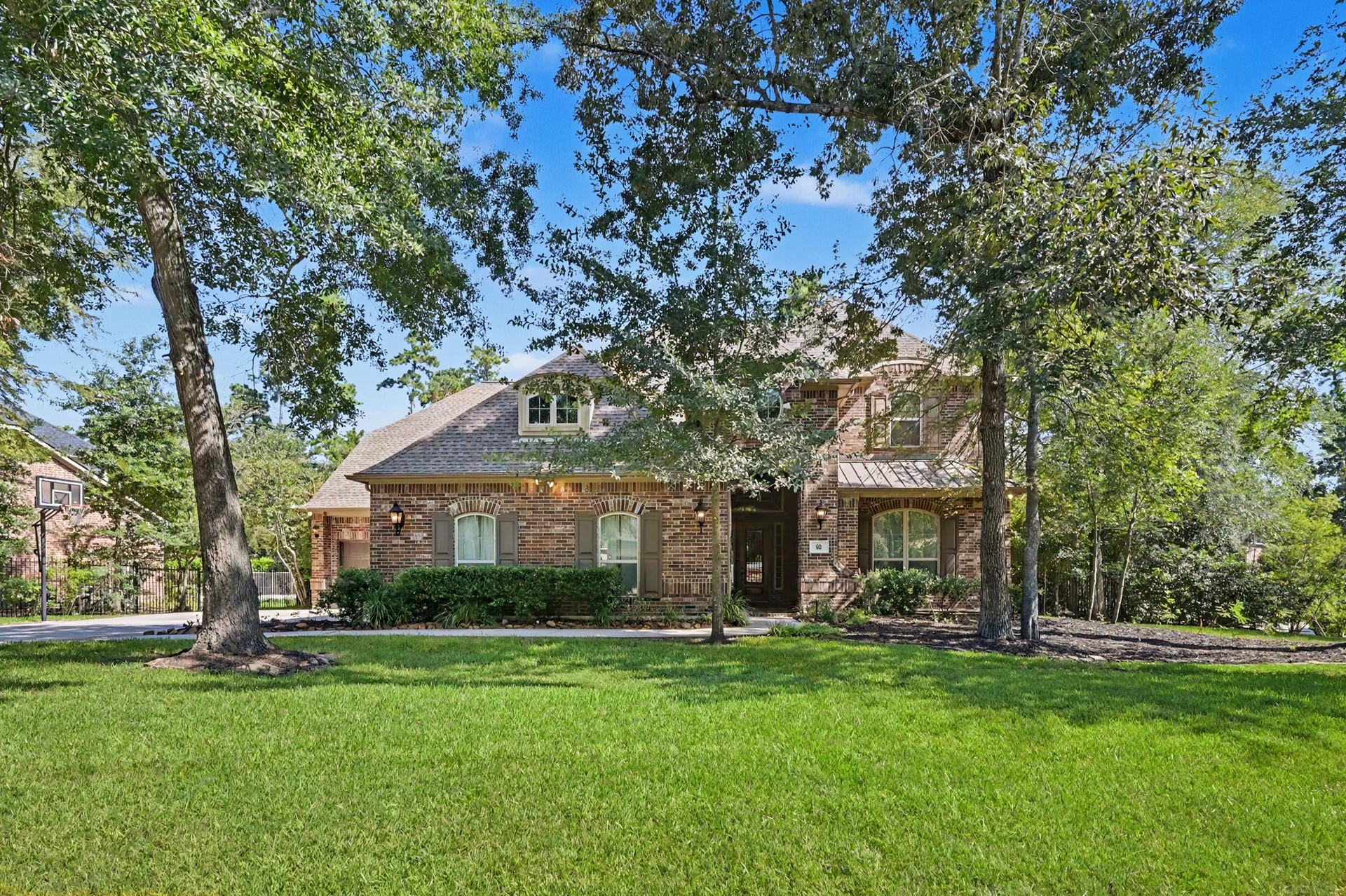 Alternate front view highlighting the sprawling lawn and mature canopy of shade trees. Features subtle architectural accents including dormer window detailing, metal roof trim, decorative shutters, and a welcoming grand entryway.