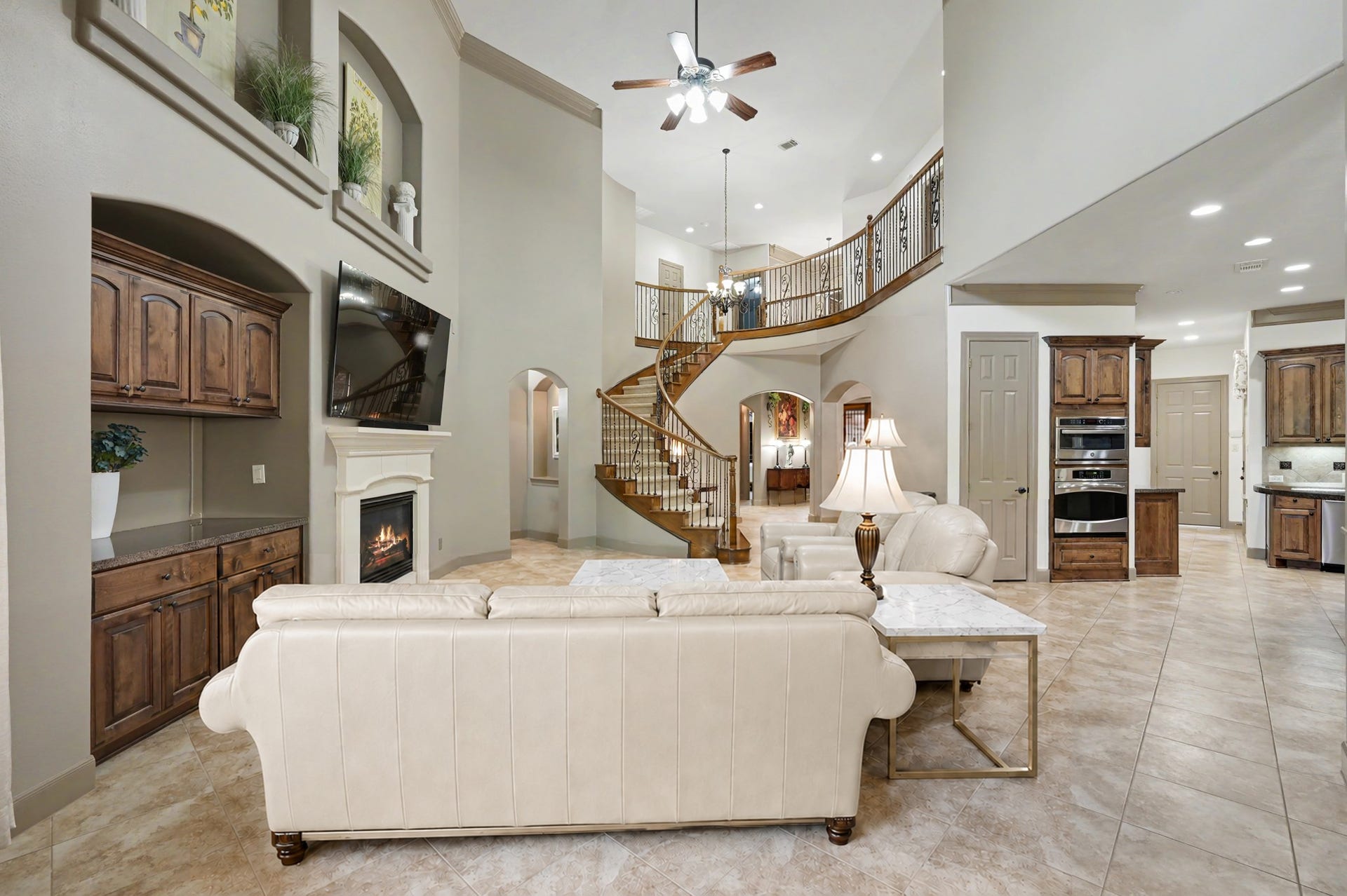 Soaring two-story family room featuring neutral tile flooring, a gas fireplace with custom built-ins, decorative art niches, and a sweeping curved staircase opening seamlessly to the kitchen. (Virtually Staged)