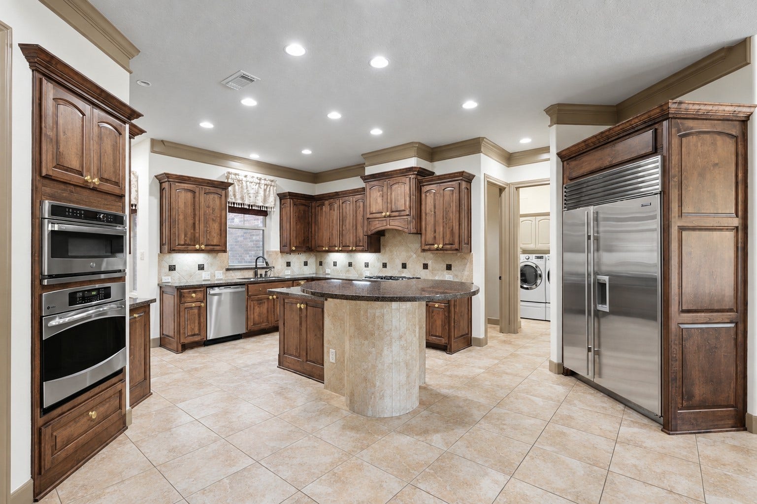 Gourmet chef's kitchen framed by rich stacked crown molding along the ceiling and custom wood cabinetry topped with matching crown trim. Highlights include granite countertops, a built-in Sub-Zero refrigerator, stainless steel double wall ovens, a central island with a tile base, tile backsplash, recessed lighting, and neutral tile flooring throughout.