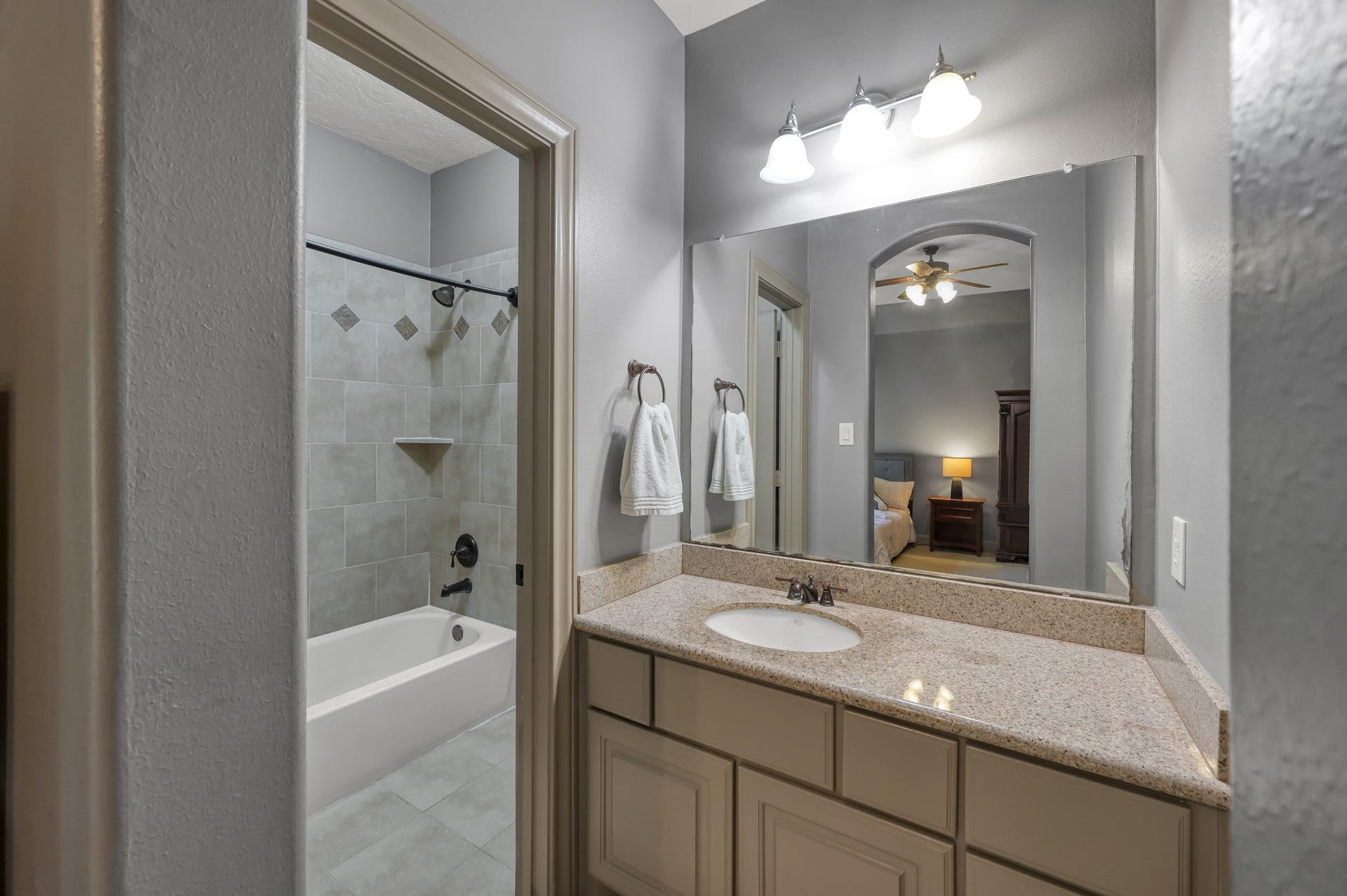 This Jack and Jill bathroom features 2 separate vanity areas with granite countertops, a large mirror, and modern lighting. The adjacent tub-shower combo has elegant tile work.