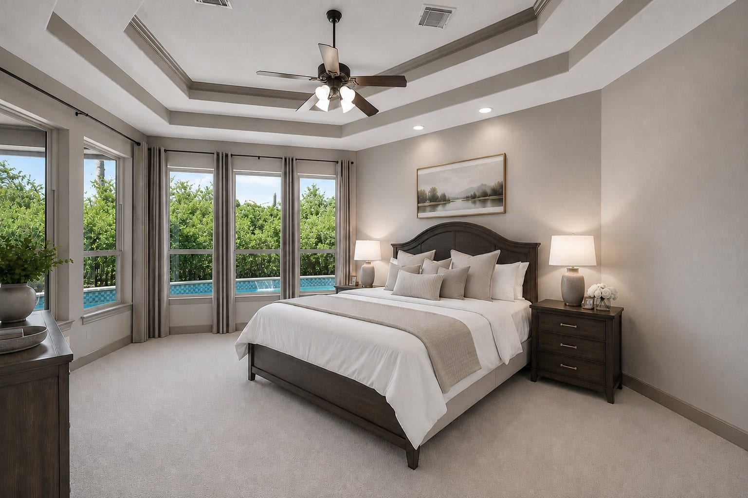 Main-floor primary retreat featuring a multi-tiered octagonal tray ceiling detailed with crown molding, a central ceiling fan, and recessed lighting. Large windows frame tranquil views of the private backyard pool, complemented by plush carpeting and fresh neutral paint. (Virtually Staged)