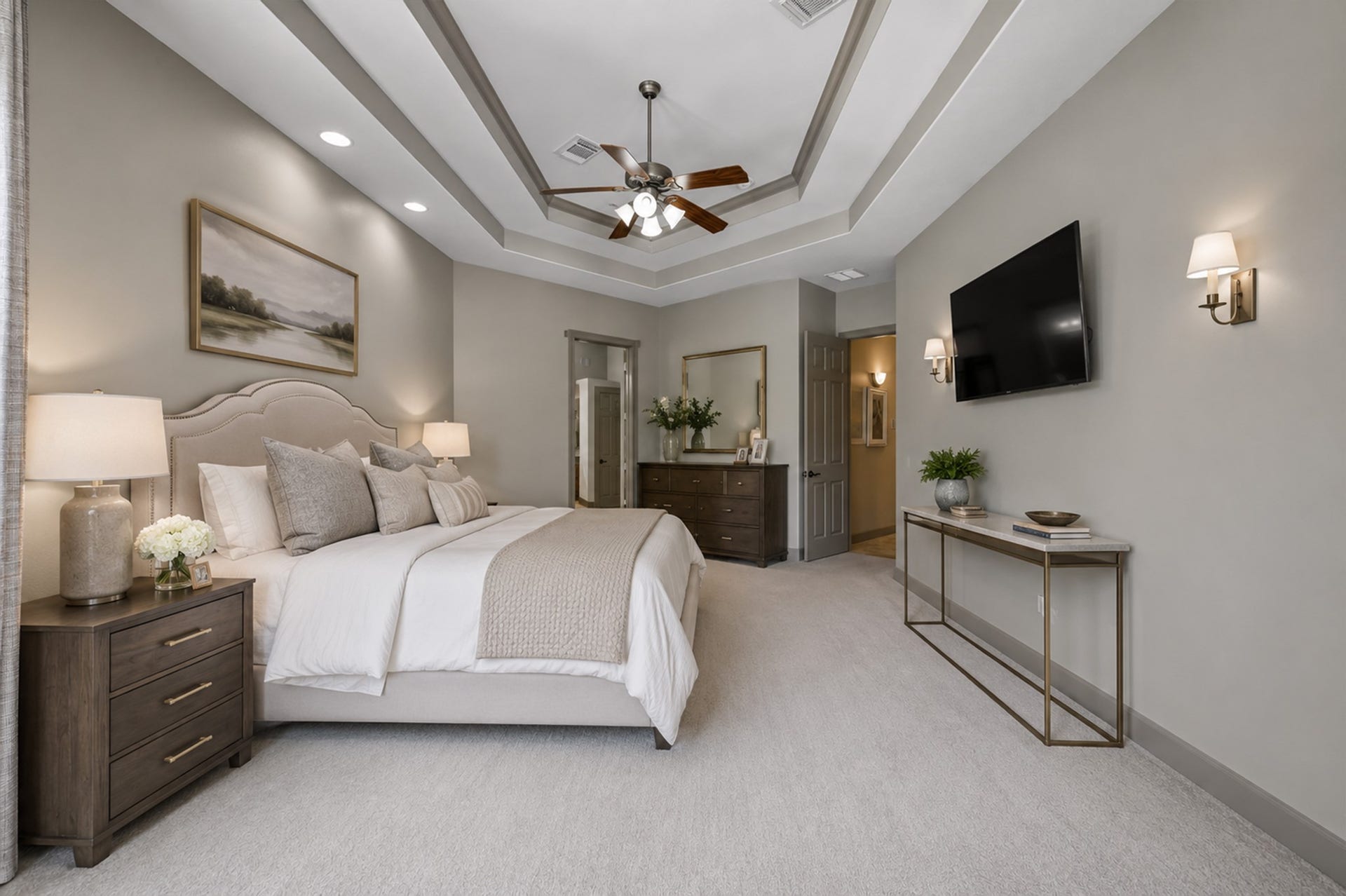 Alternate view of the main-floor primary retreat showcasing the multi-tiered tray ceiling with custom accent molding, central ceiling fan, and dedicated accent recessed lighting. Finished with plush carpeting, fresh paint, and direct access to the ensuite bath. (Virtually Staged)