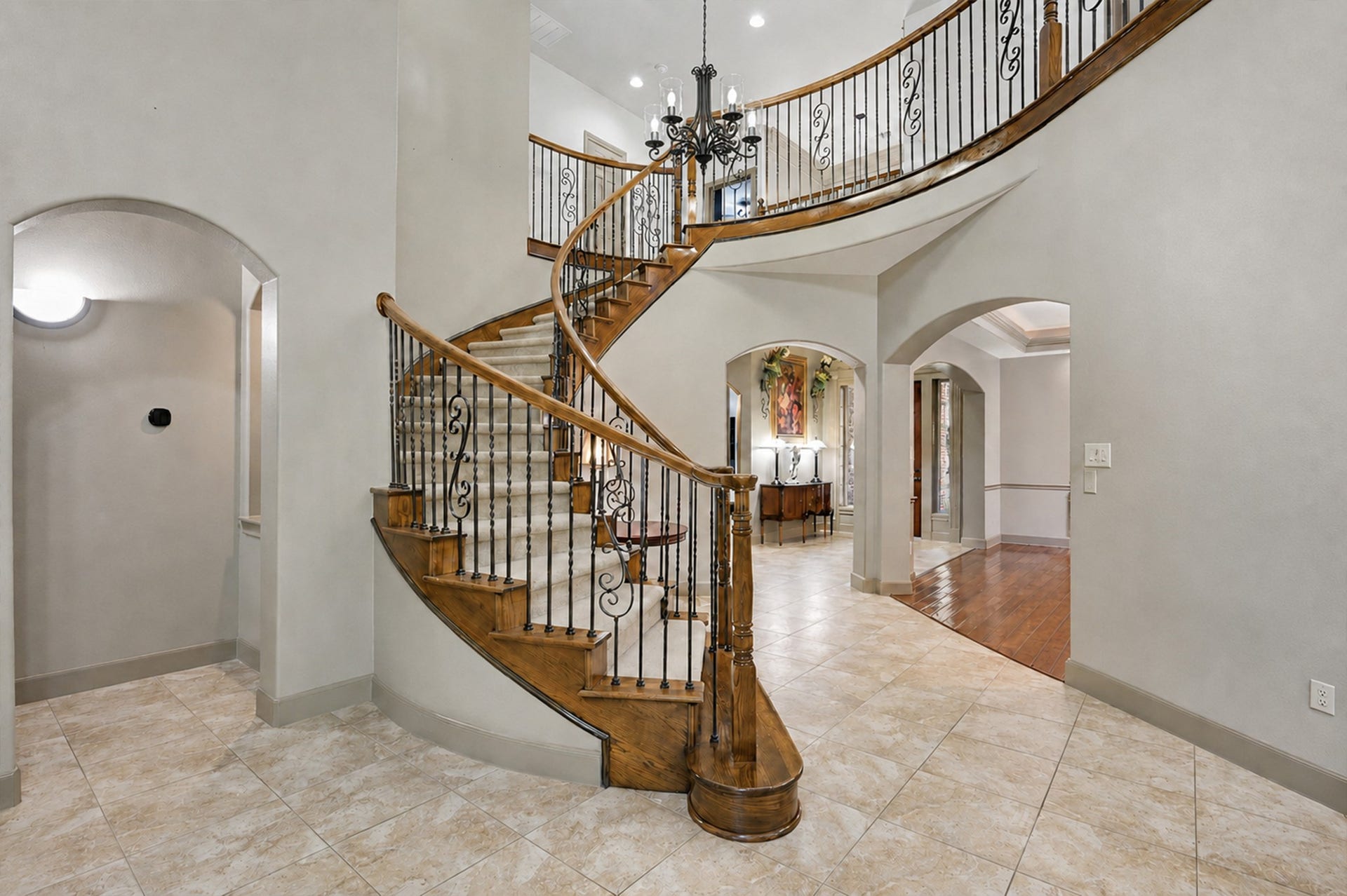 This elegant home features a grand curved staircase with decorative metal pickets, a chandelier, graceful archways, and tile floors, all leading into a warm, inviting living space