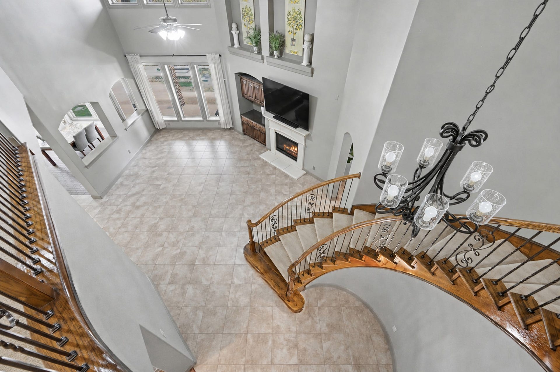 Overhead perspective looking down from the catwalk, offering a clear visual representation of the main floor's true scale and footprint. Showcases the sweeping staircase with wrought-iron balusters, dramatic chandelier, tile flooring, gas fireplace, and light-filled pool views.