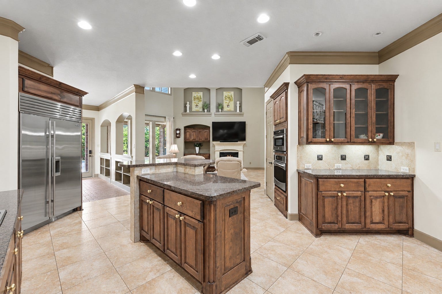 Seamless open-concept view from the kitchen showing rich custom cabinetry topped with crown molding, granite countertops, and neutral tile flooring throughout. Features a built-in buffet with glass-front display cabinets, under-cabinet lighting, a central island with dual-level breakfast bar counter, recessed lighting, and a built-in Sub-Zero refrigerator.
