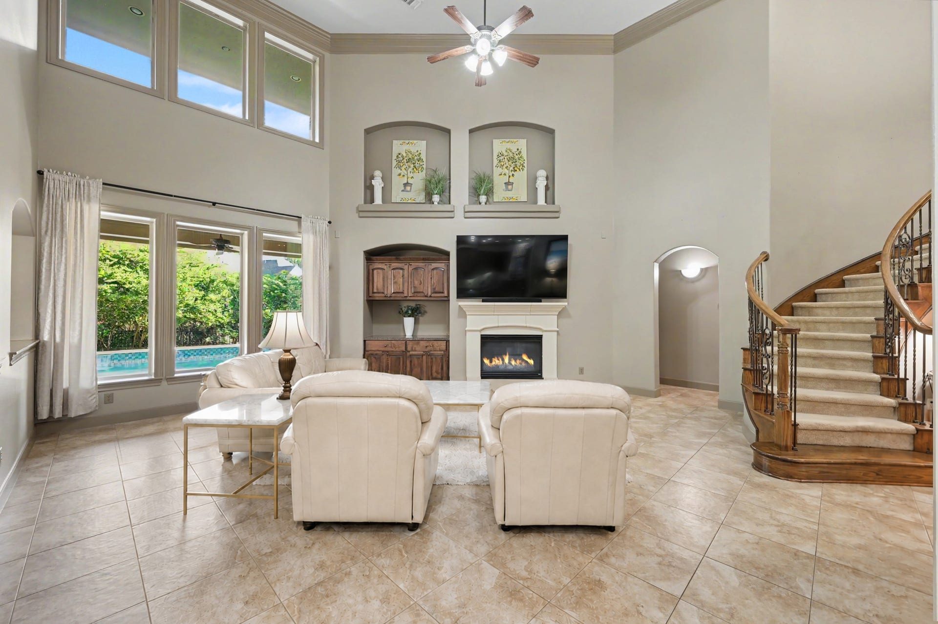 Bright two-story family room with tile floors, a stone fireplace, built-in cabinetry, display niches, and floor-to-ceiling pool views framed by a grand curved staircase. (Virtually Staged)