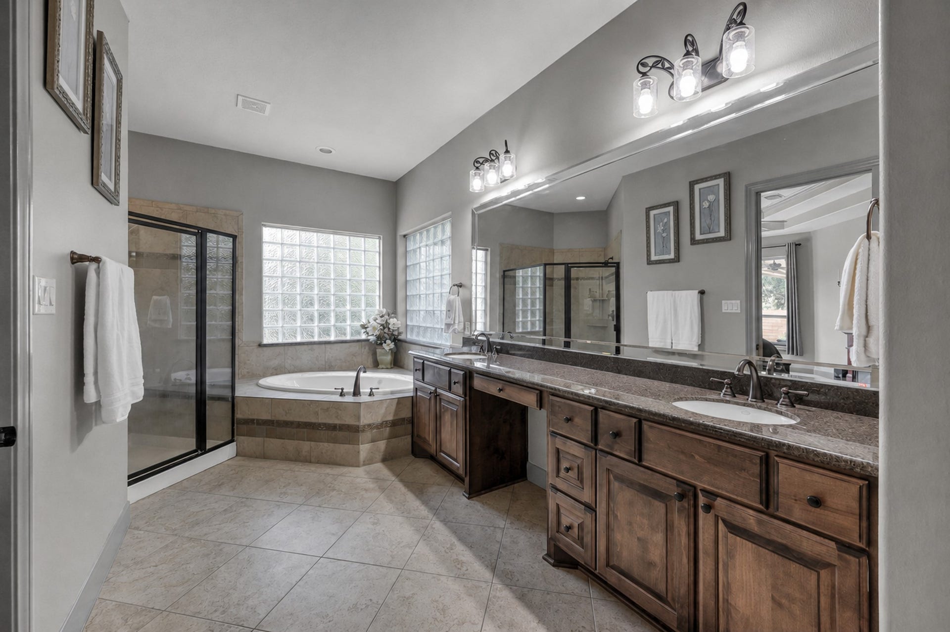 This Oversized primary bathroom features a double long vanity with dual sinks, a corner jacuzzi tub, and a separate glass-enclosed shower. It has elegant tile flooring, ample natural light through glass block windows, and stylish lighting fixtures above the mirror.