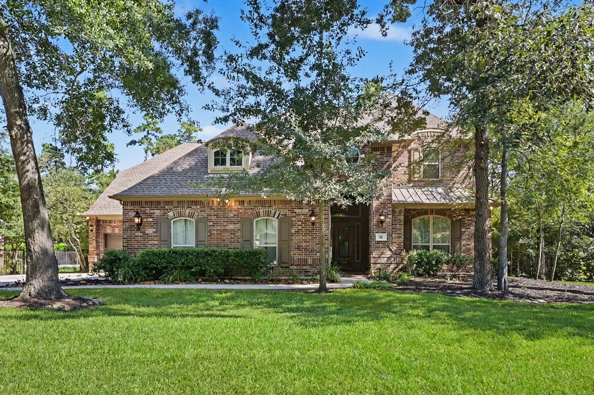 Front view highlighting the sprawling lawn and mature canopy of shade trees. Features subtle architectural accents including dormer window detailing, metal roof trim, decorative shutters, and a welcoming grand entryway.
