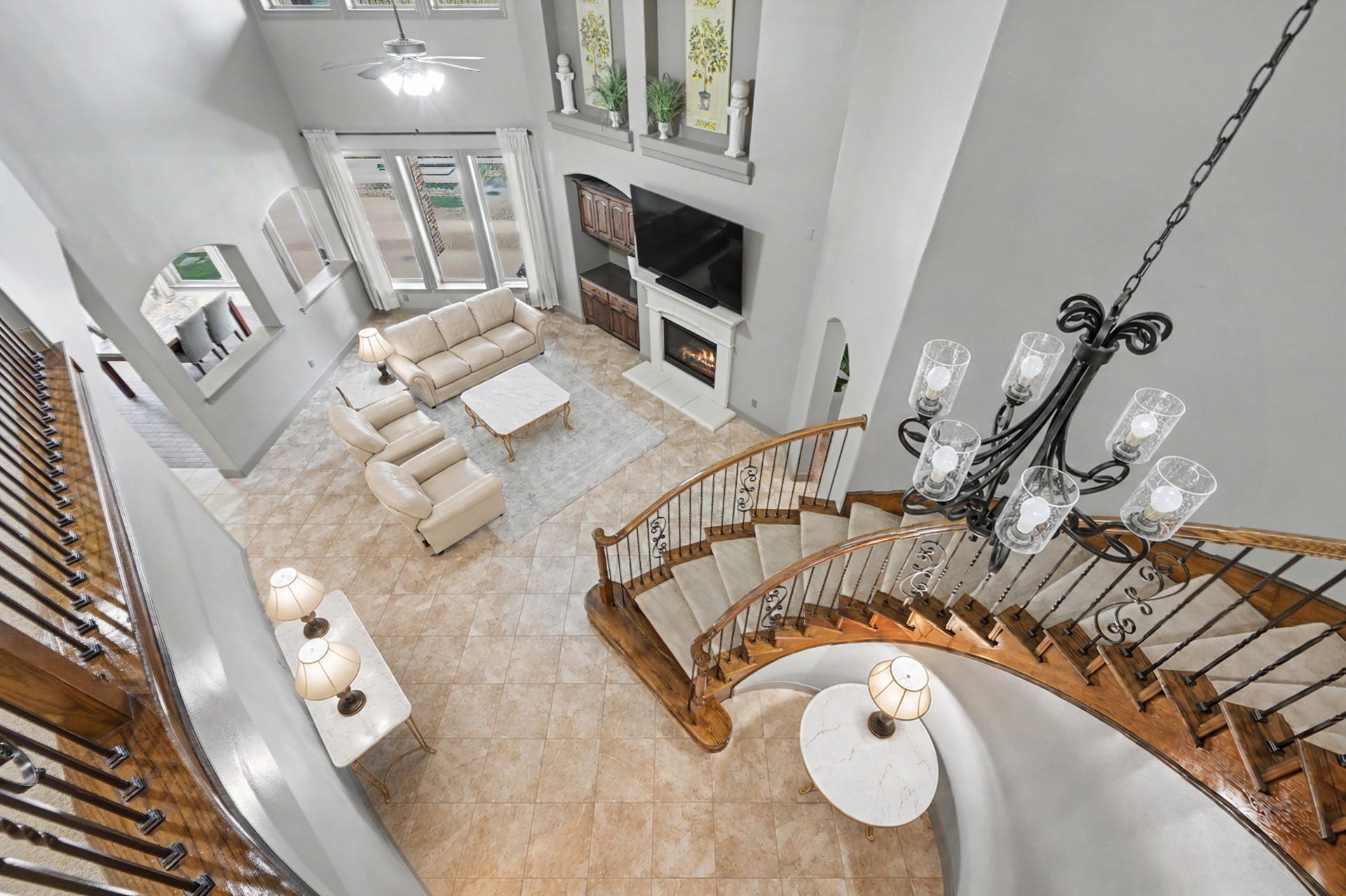 View from the second-story catwalk overlooking the family room, highlighting the grand curved staircase, stone fireplace, high ceilings, and floor-to-ceiling windows. (Virtually Staged)