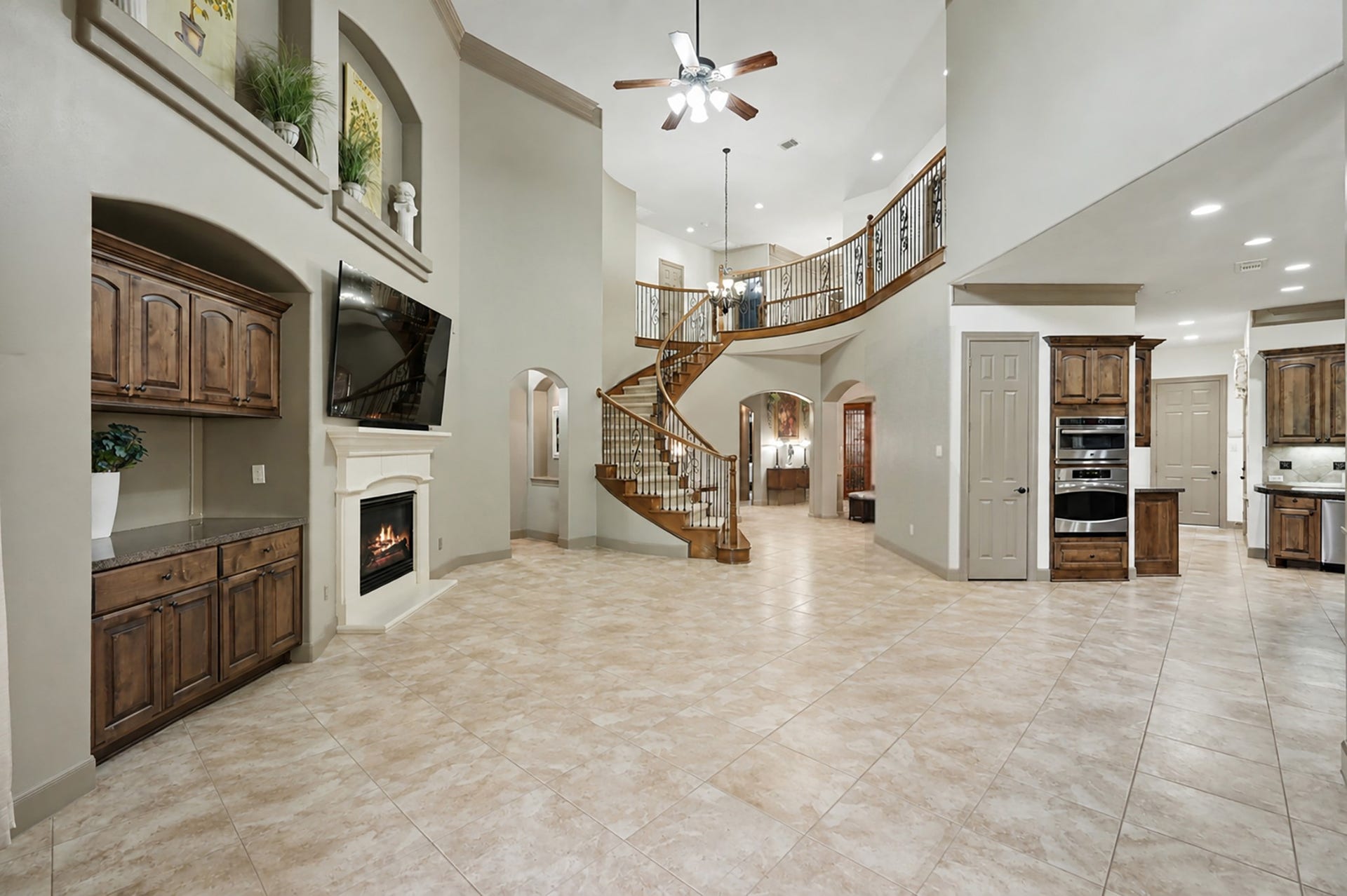 An unfurnished perspective providing a better visual representation of how the living space flows effortlessly into the kitchen and foyer. Highlights the soaring ceilings, built-in cabinetry, double ovens, and sweeping curved staircase.