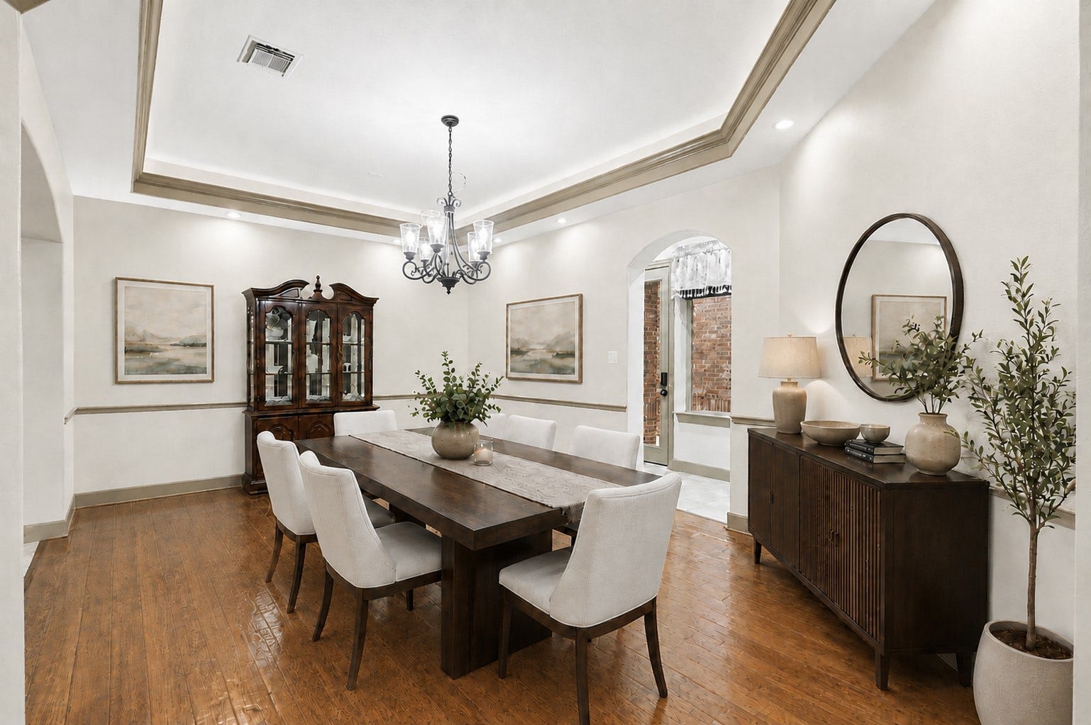 Formal dining space showcasing warm wood floors and refined double-tiered tray ceiling with crown molding and subtle tray lighting. Finished with a central chandelier, recessed lights, chair rail trim, and graceful arched pass-throughs. (Virtually Staged)