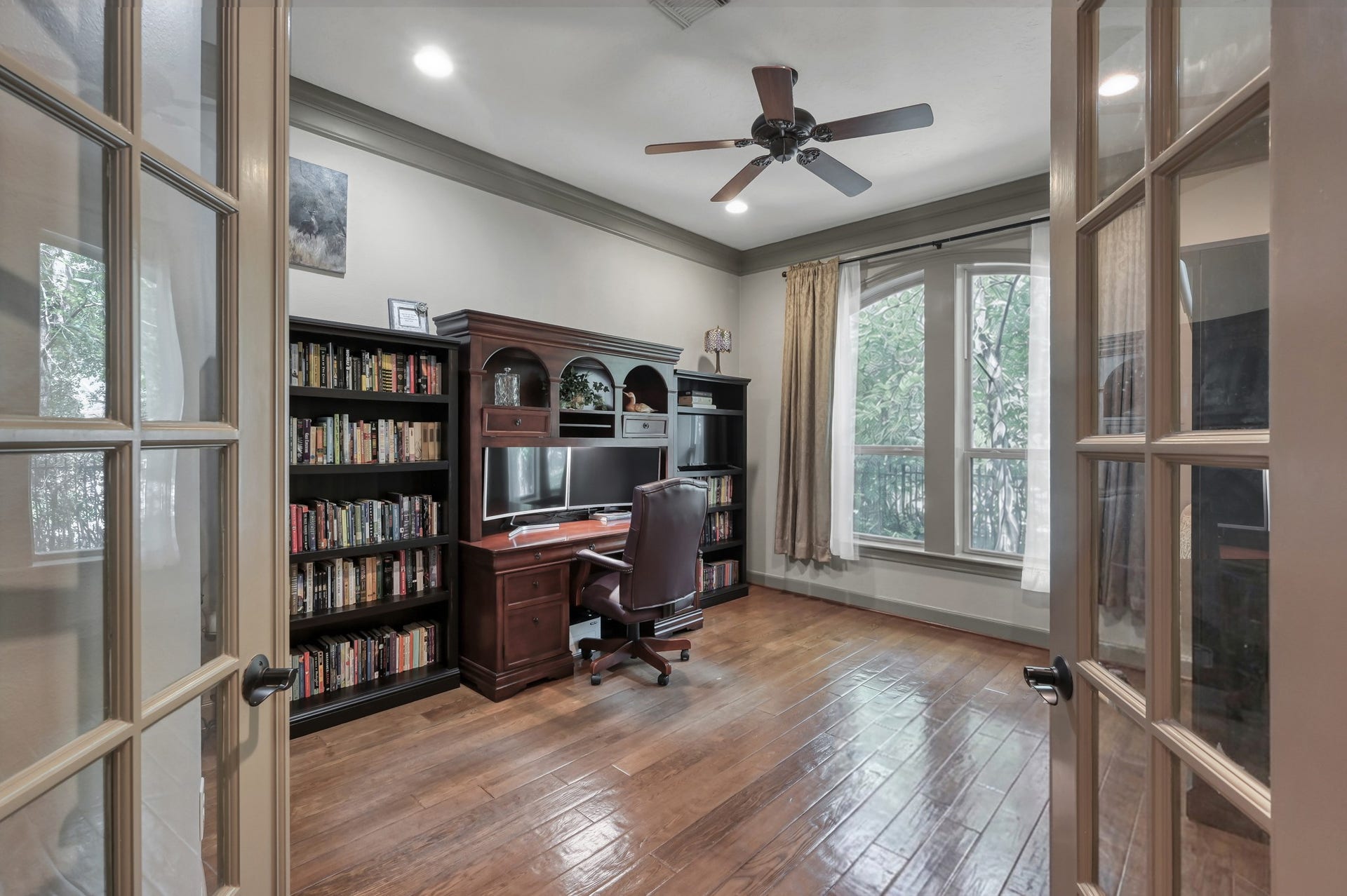 Private home office entered through glass double French doors. Features rich hand-scraped hardwood flooring, continuous dark crown molding, recessed lighting, a ceiling fan, and large arched windows that fill the space with natural light and peaceful views.