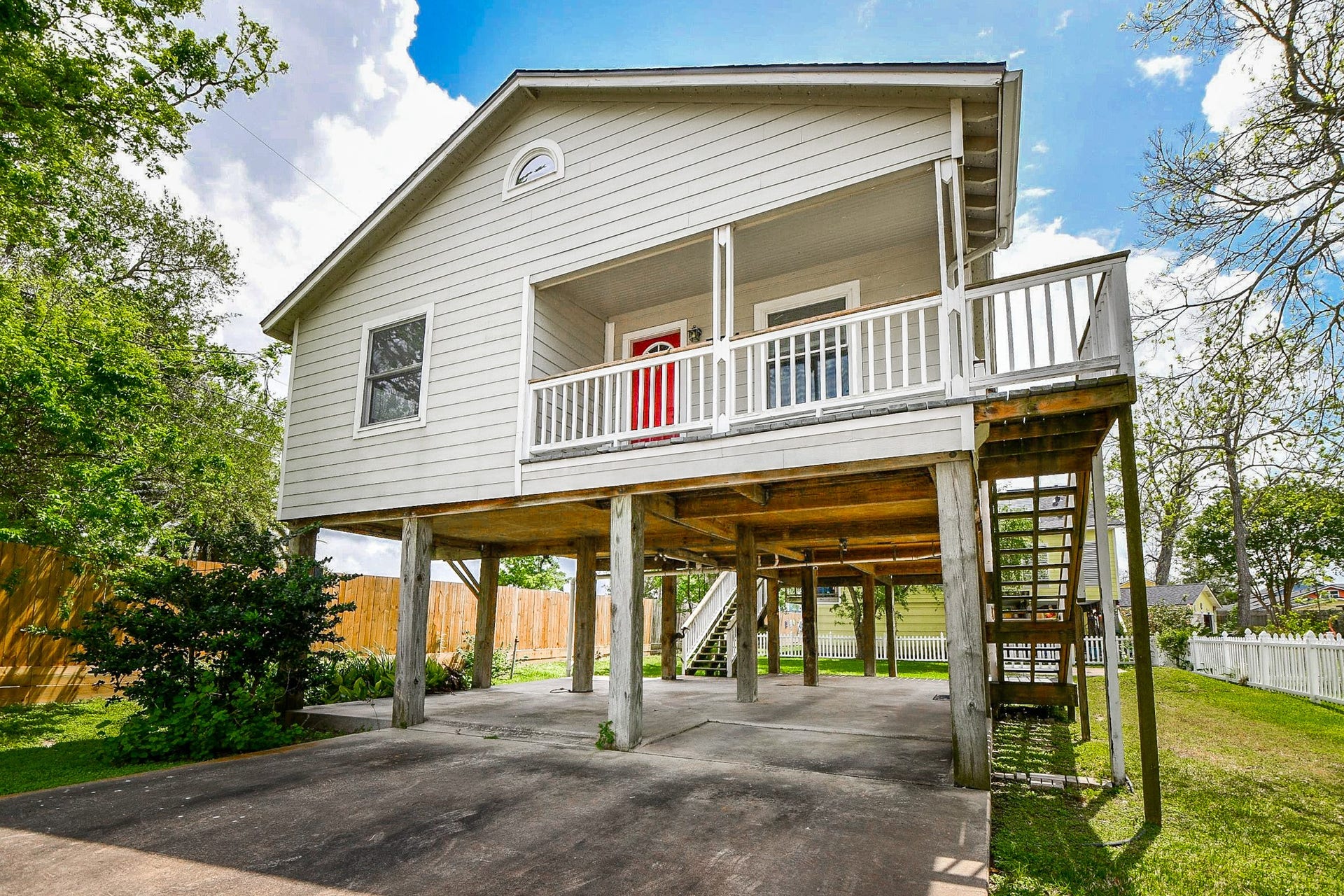 704 6th Street, Kemah, TX 77565 Elevated home with a charming front porch and red door, featuring an open carport underneath. Surrounded by a fenced yard with mature trees, offering a blend of privacy and space.