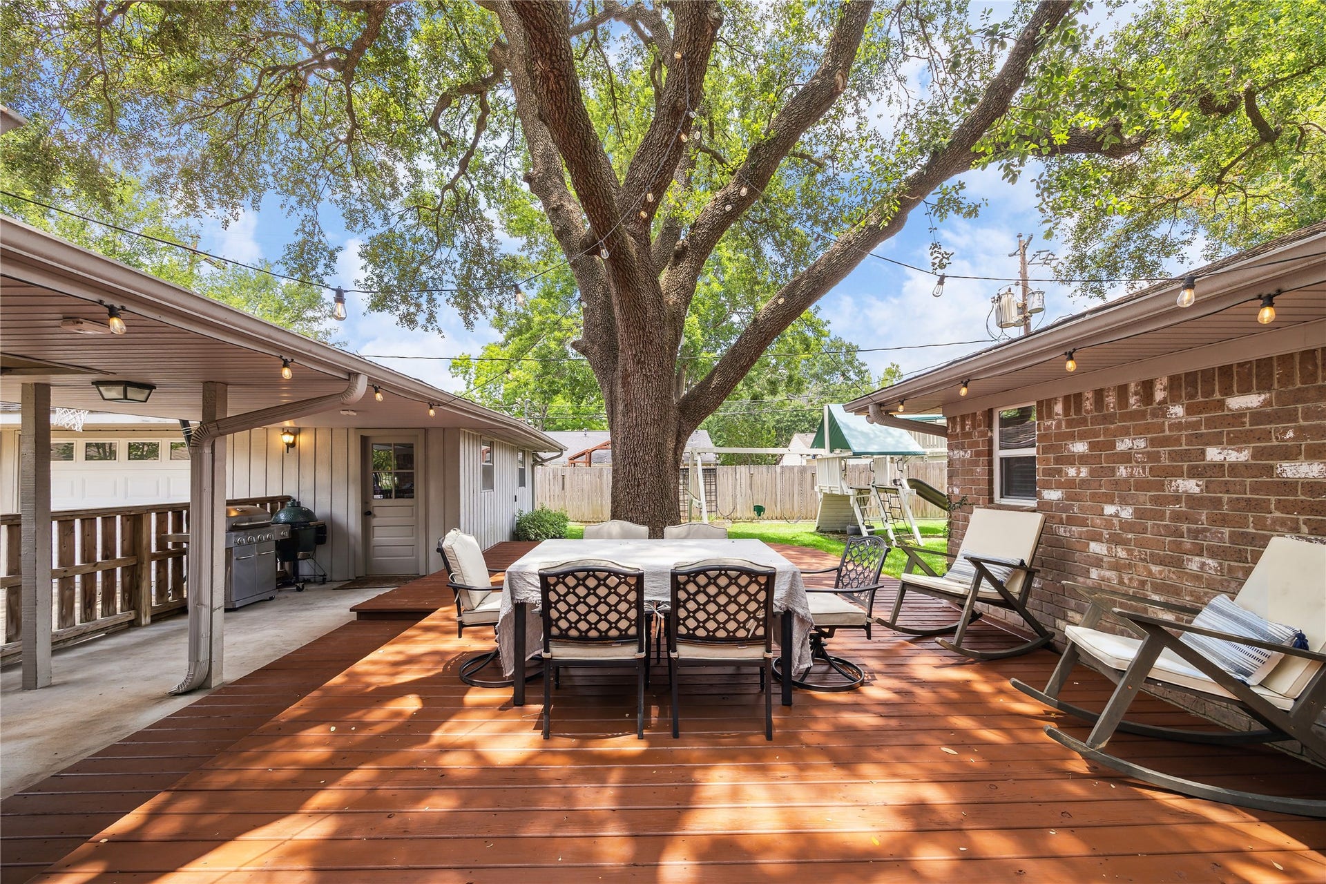 10714 Piping Rock Lane, Houston, TX 77042 A spacious wood deck beneath a mature tree offers room for outdoor dining and lounging, with string lights overhead and a covered area alongside.