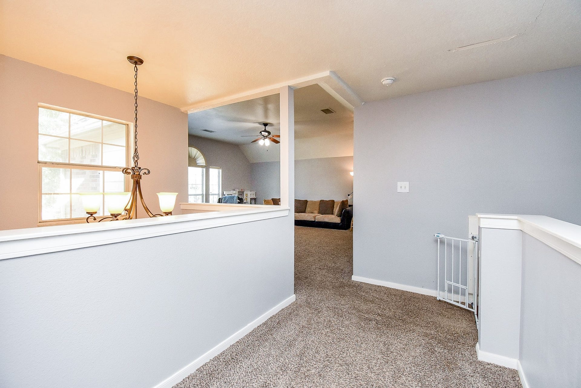 6703 Morning Glory Trace, Richmond, TX 77407 This photo shows a bright upstairs hallway with carpeted flooring and soft gray walls. A decorative light fixture hangs near a window, and the space opens into a cozy living area with a ceiling fan and large sectional sofa.