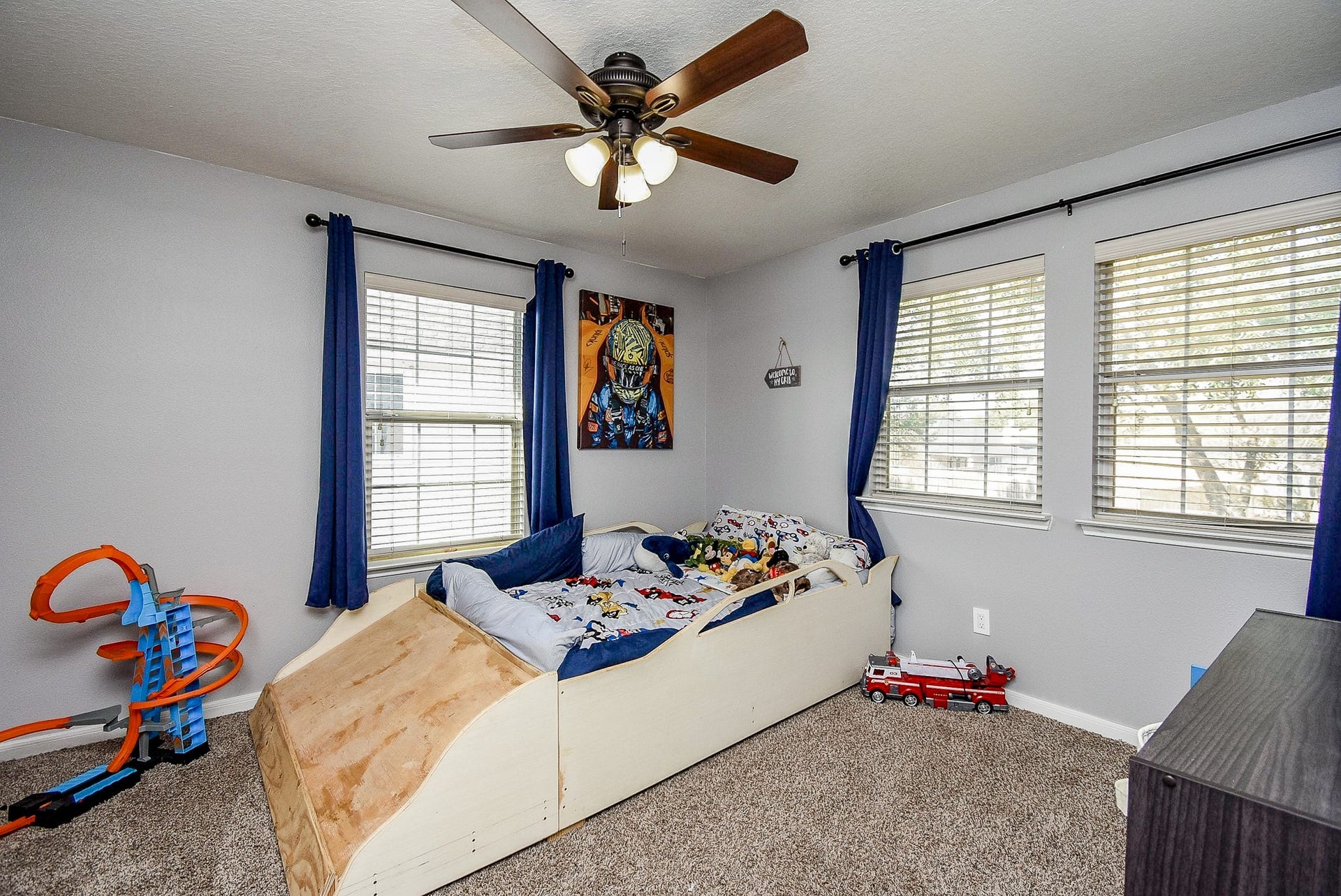 6703 Morning Glory Trace, Richmond, TX 77407 Cozy children's bedroom with carpeted flooring, The room has ample natural light from three windows, accented by blue curtains. A ceiling fan ensures comfort, while the neutral walls provide a calming backdrop. Perfect for a child's personal space.