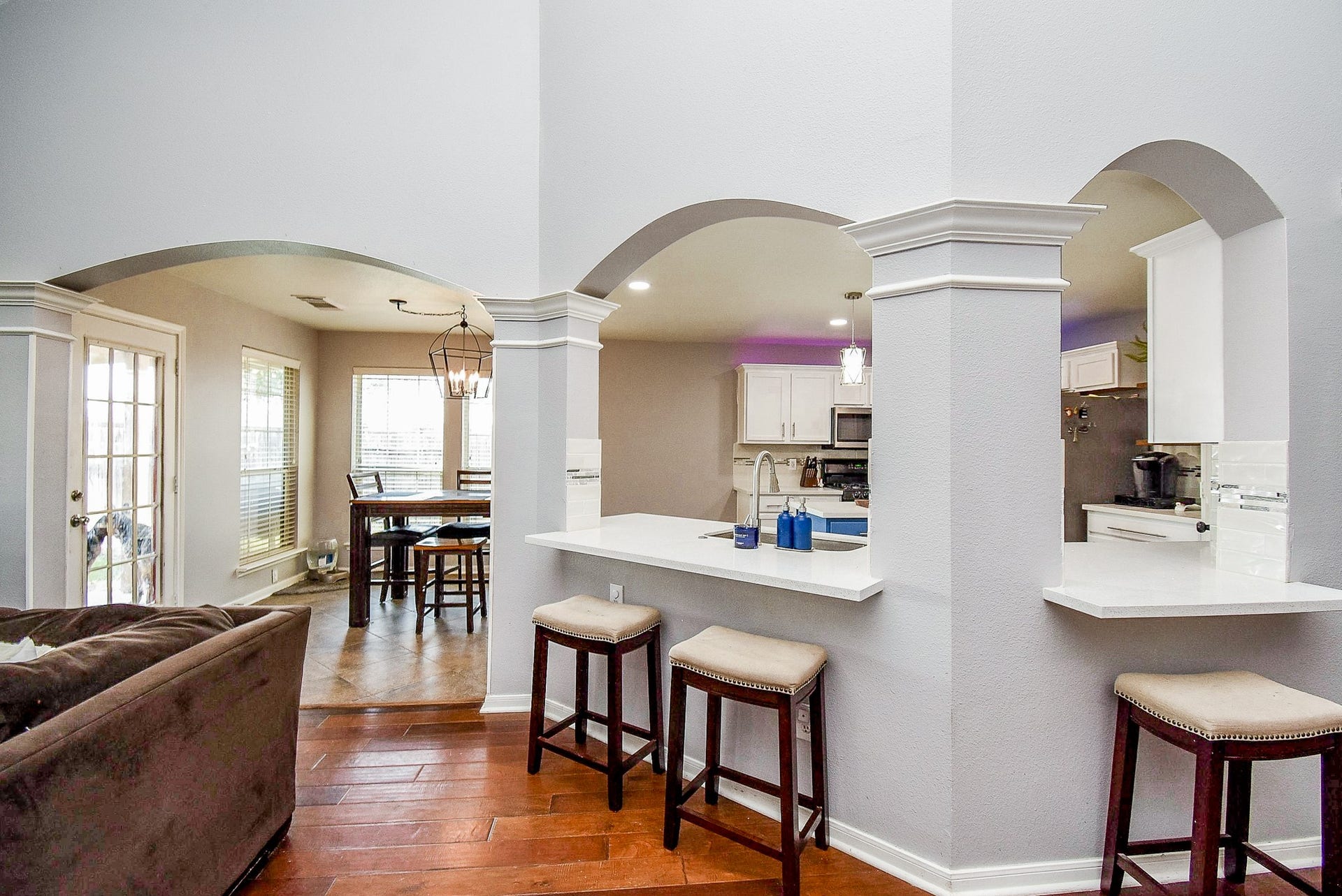 6703 Morning Glory Trace, Richmond, TX 77407 This photo showcases a bright, open-concept living space with arched entryways. The kitchen features modern white cabinetry and a breakfast bar with seating. Adjacent is a cozy dining area with large windows and abundant natural light, complemented by warm hardwood floors.