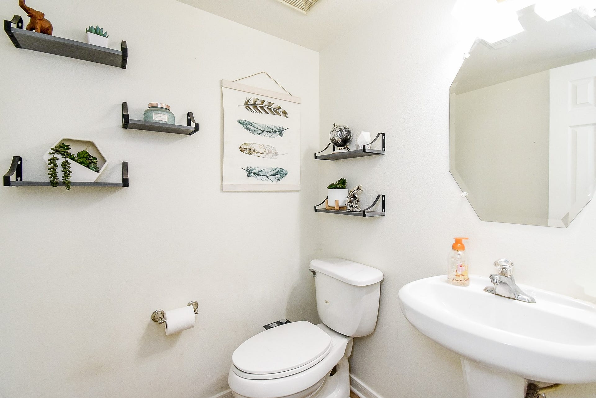 6703 Morning Glory Trace, Richmond, TX 77407 This is a compact, modern bathroom featuring a toilet and pedestal sink. It has wall-mounted shelves with decorative items and artwork, creating a clean and stylish look. The space is well-lit, offering a fresh and inviting atmosphere.