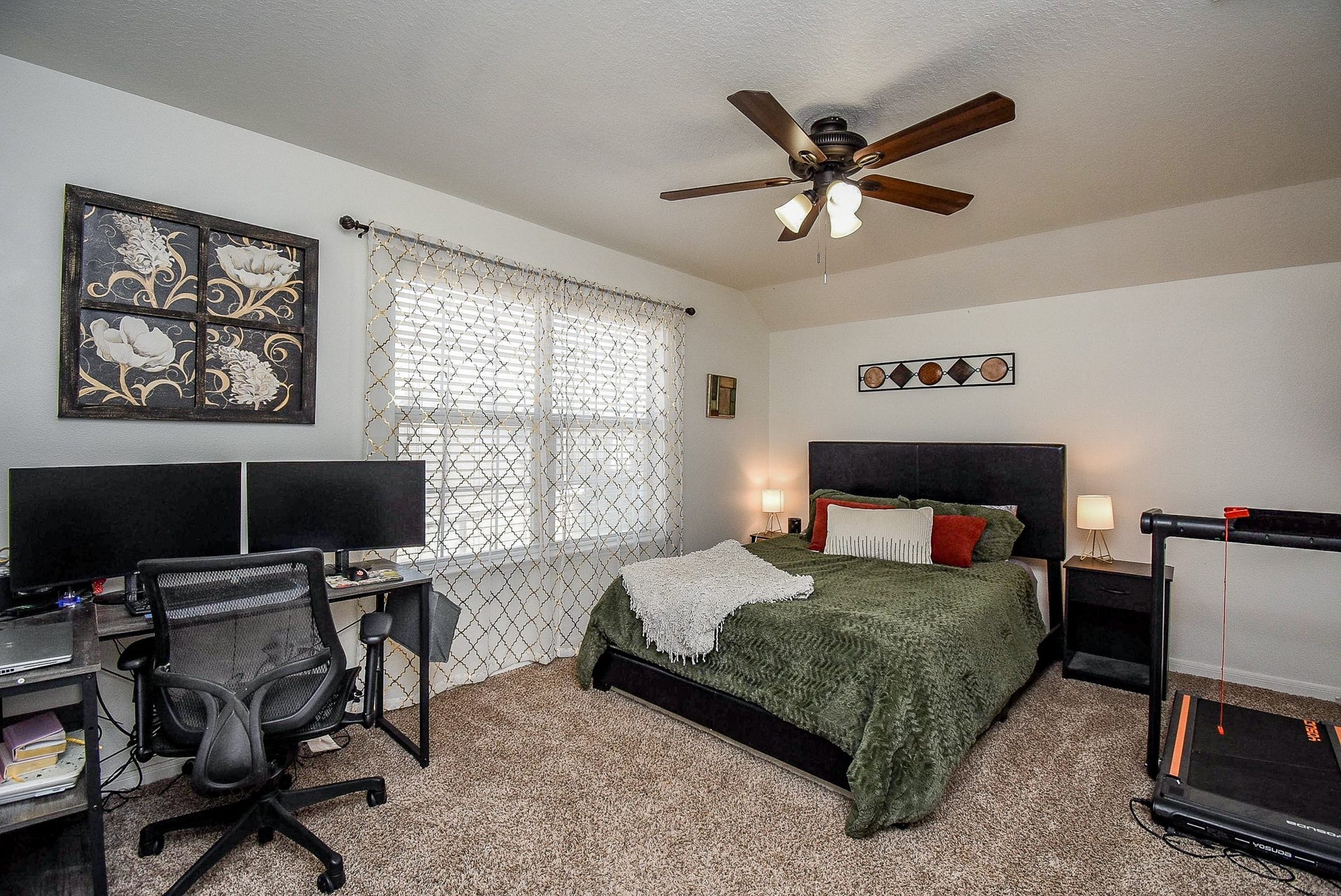 6703 Morning Glory Trace, Richmond, TX 77407 Cozy bedroom with a large bed, stylish decor, and a ceiling fan. Features a home office setup with dual monitors and a treadmill. Bright and inviting with natural light from a window.