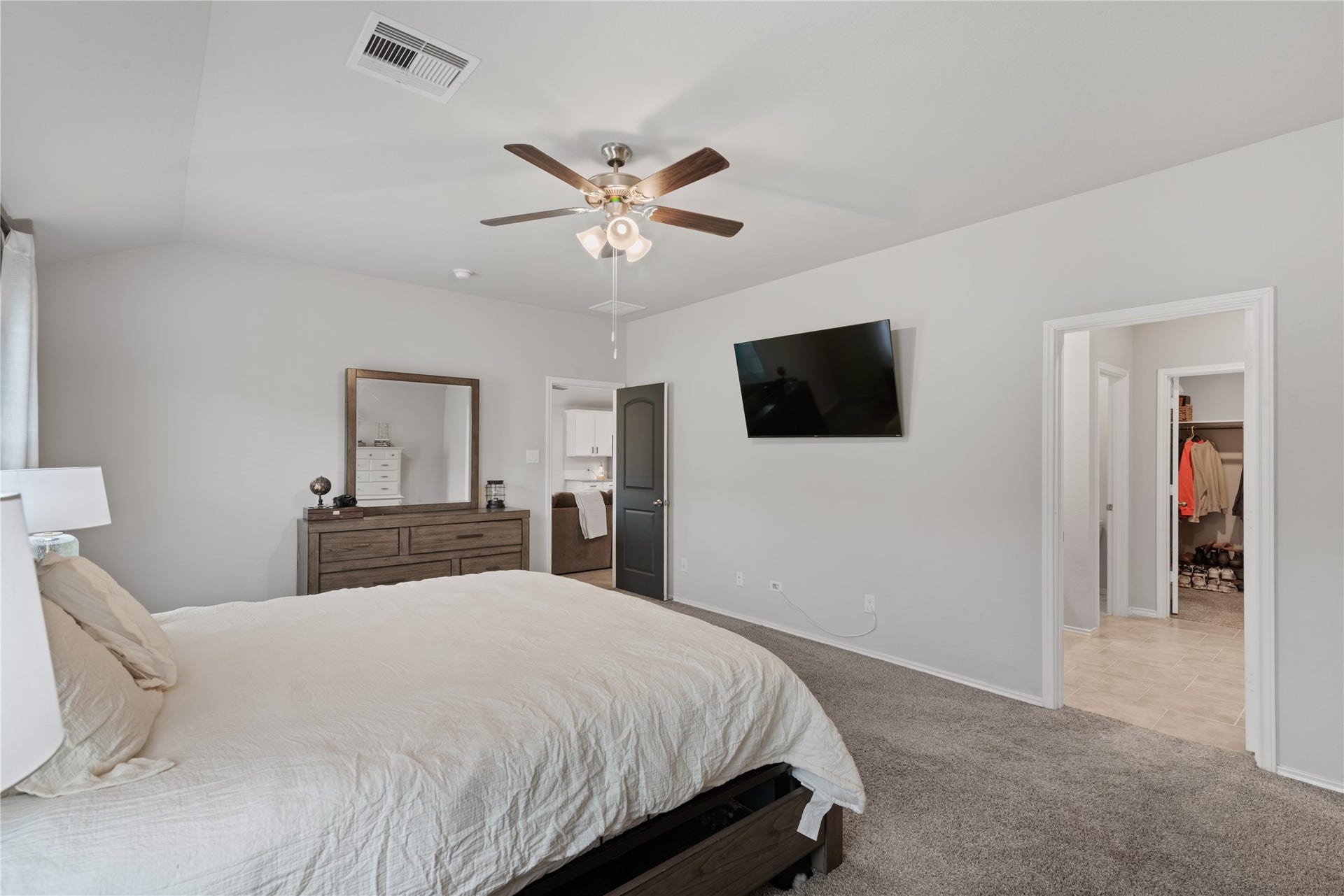 Spacious bedroom with a cozy carpet, featuring a ceiling fan and wall-mounted TV for relaxation. The room includes a well-organized walk-in closet and access to a modern bathroom, enhancing convenience and comfort.