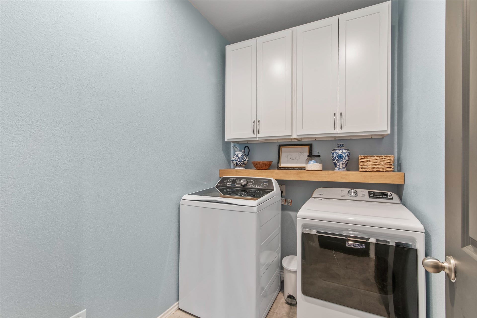 Charming laundry room with modern washer and dryer, ample storage in white cabinetry, and a convenient wooden shelf for decor and essentials.