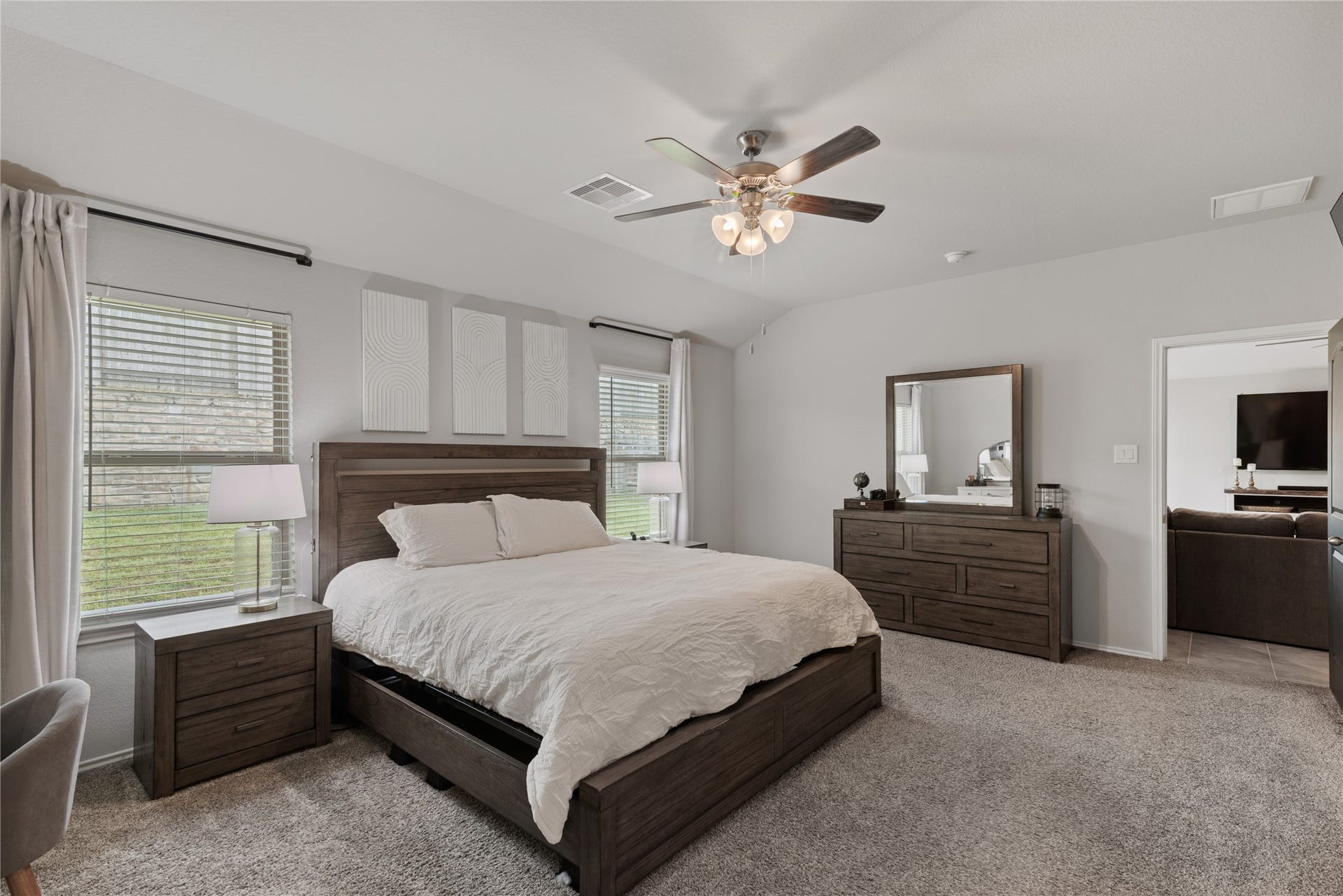 Spacious and inviting master bedroom featuring a cozy carpeted floor, elegant ceiling fan, and ample natural light from dual windows. The room includes a stylish wood bed frame, matching nightstands, and a dresser with a large mirror, exuding comfort and modern charm.