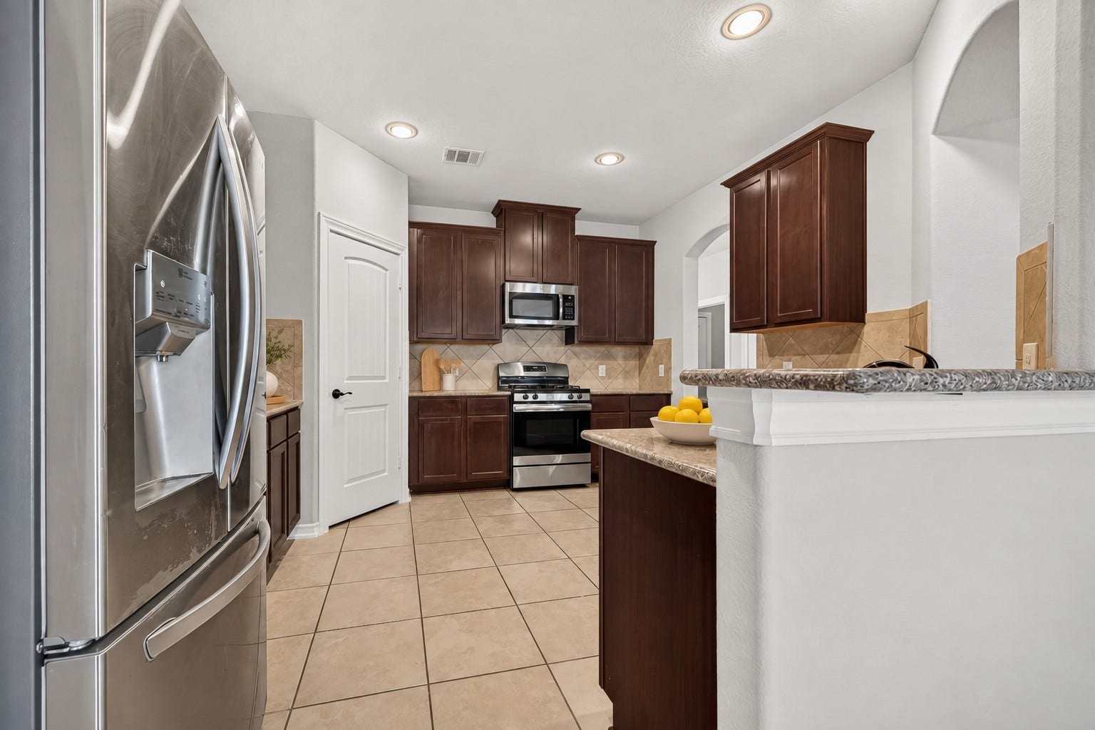 3658 Diamond Creek Drive, Missouri City, TX 77459 Kitchen features rich wood cabinetry, tile flooring, and stainless steel appliances. Interior opening above the sink provides a view into the living area.