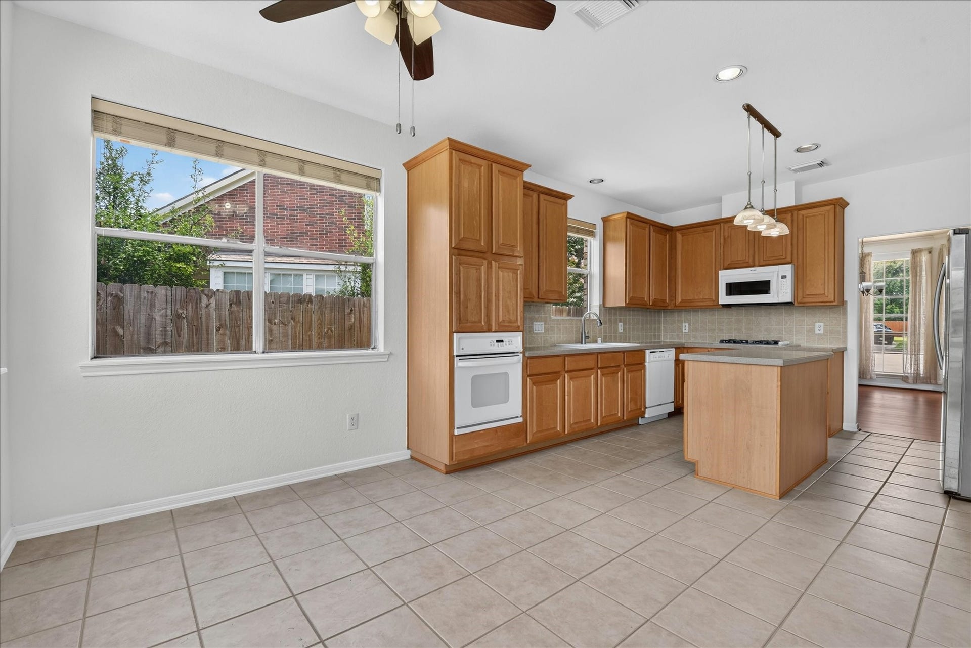 4610 Misty Morning Court, Missouri City, TX 77459 This photo showcases a spacious kitchen with wooden cabinets, a central island, and modern appliances. It features tiled flooring, ample natural light from large windows, and a ceiling fan, creating a bright and welcoming atmosphere.