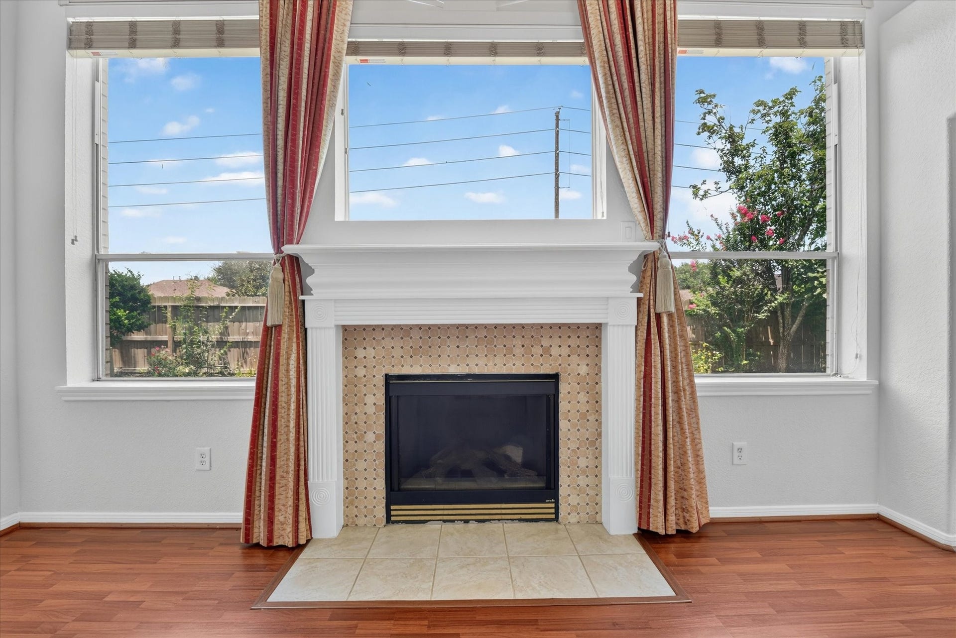 4610 Misty Morning Court, Missouri City, TX 77459 The photo features a cozy living room with a central fireplace framed by elegant red and gold curtains. Large windows on either side offer plenty of natural light and views of the backyard, creating a warm and inviting atmosphere. The room has polished wood flooring, adding to its charm.