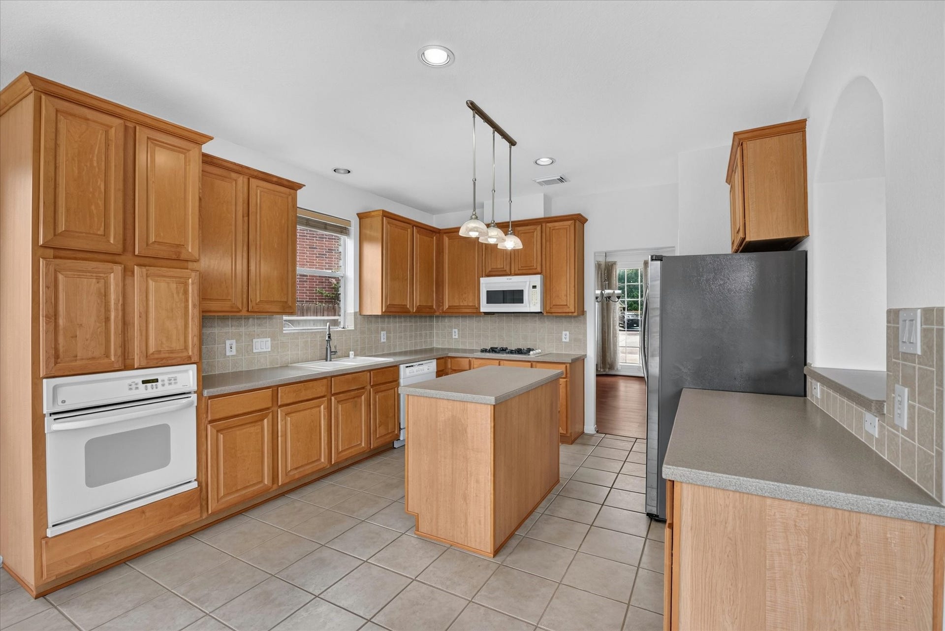 4610 Misty Morning Court, Missouri City, TX 77459 This spacious kitchen features wooden cabinetry, tiled flooring, and modern appliances including a built-in oven and microwave. It has a central island, ample counter space, and natural light from a window. The room opens to another area through a glass door.