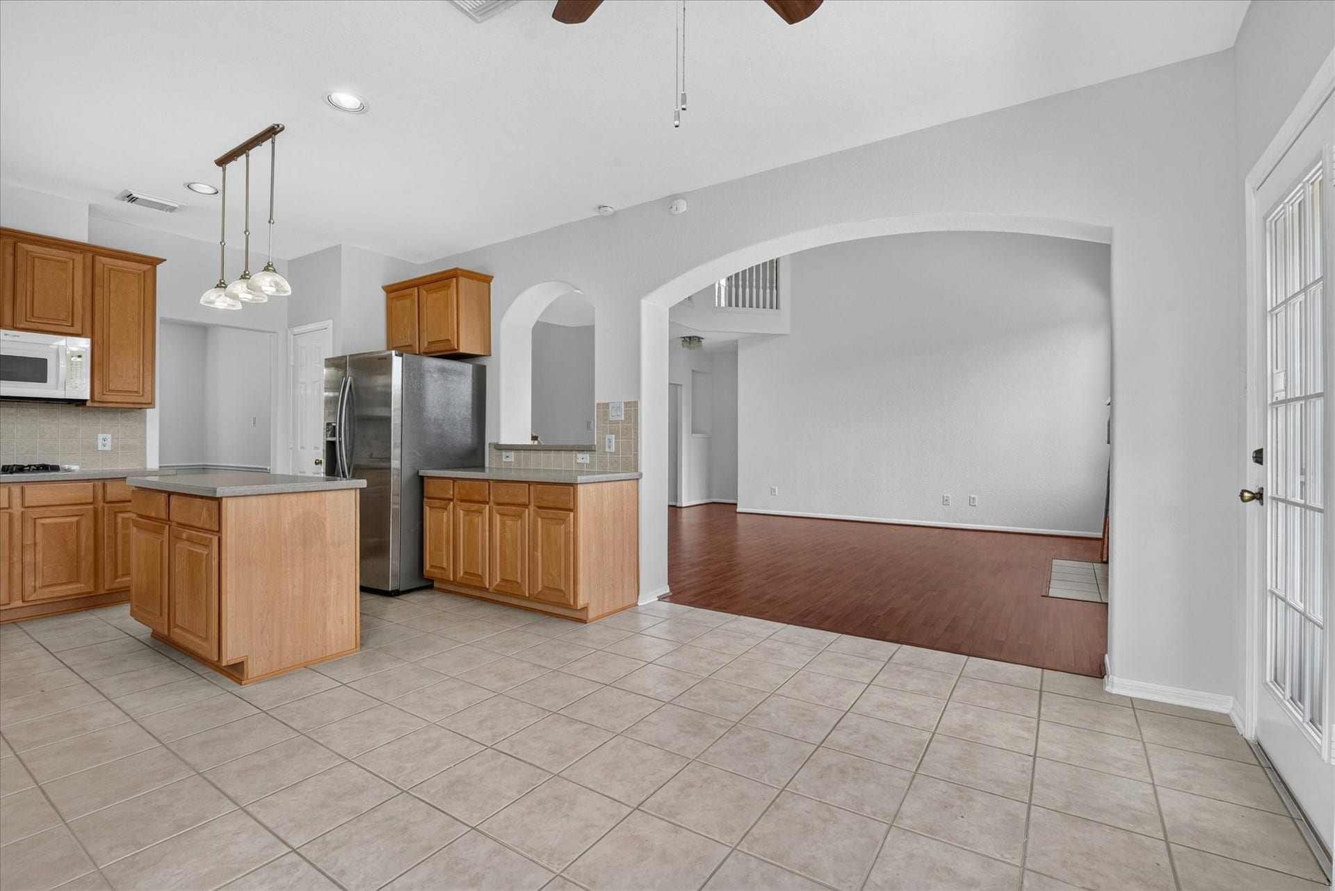 4610 Misty Morning Court, Missouri City, TX 77459 This photo showcases a spacious open-concept kitchen and living area. The kitchen features wooden cabinetry, a modern stainless steel refrigerator, and an island with pendant lighting. It opens to a living space with hardwood floors, visible through an elegant archway, enhancing the home's airy feel.