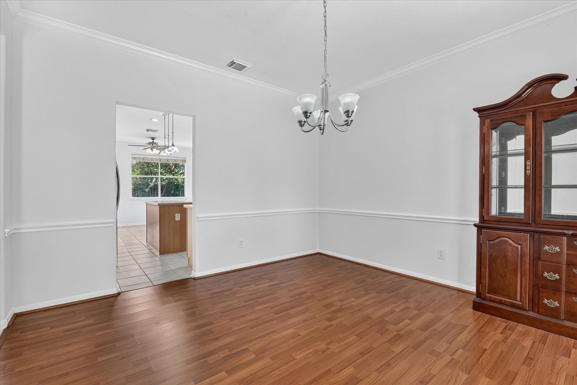 4610 Misty Morning Court, Missouri City, TX 77459 This photo shows a bright dining room with wood laminate flooring, a classic chandelier, and a glass-fronted cabinet. It opens into a kitchen with a window providing natural light.