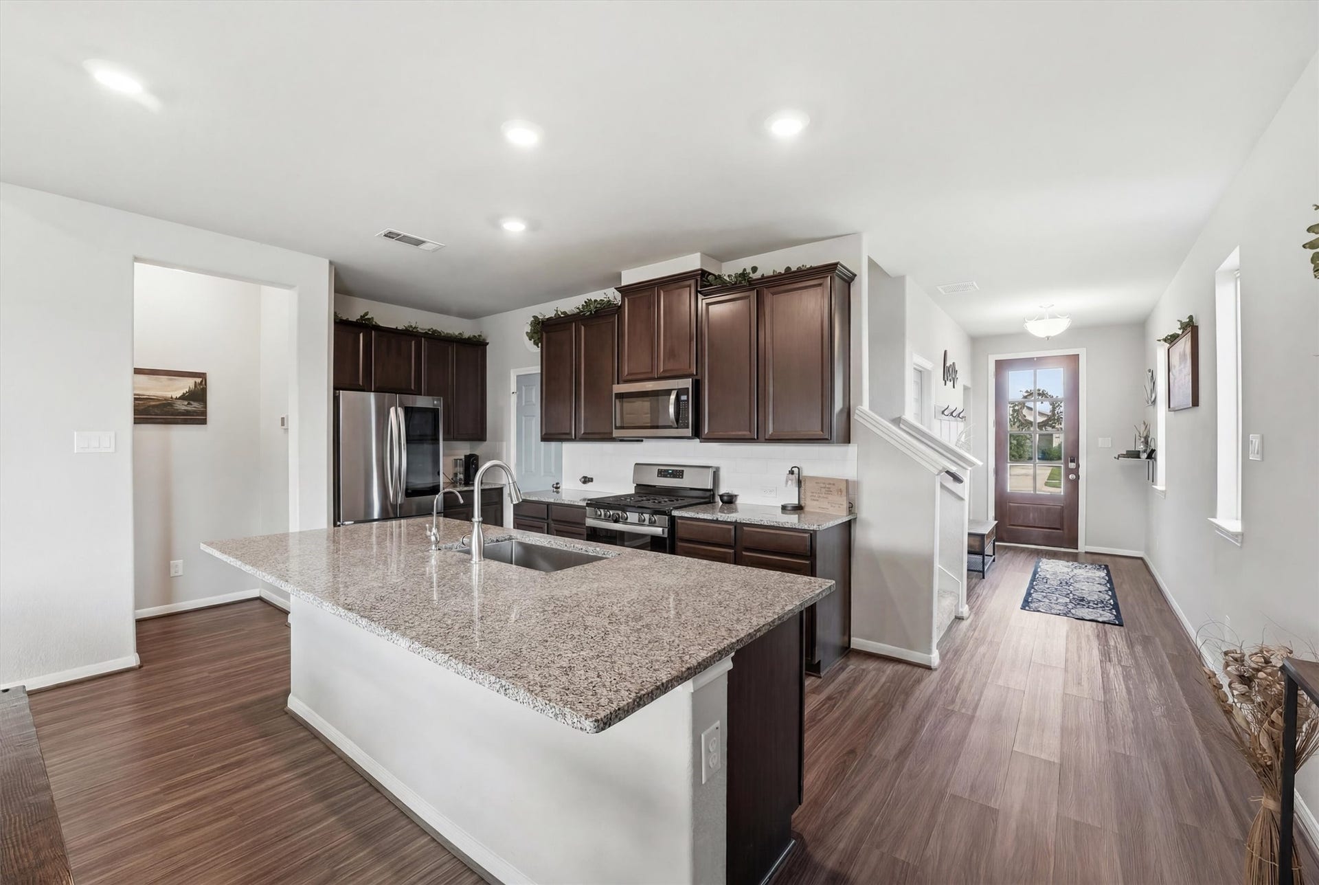 2130 Shellbark Way, La Marque, TX 77568 This modern kitchen includes a large island with a sink, stainless steel appliances, and dark wood cabinets. The open layout leads to an entryway with a wooden floor and a glass-paneled front door, creating a bright and inviting space.