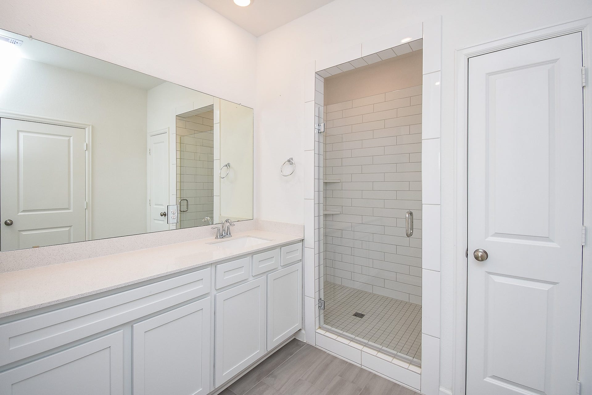 A modern primary bathroom featuring a large vanity with ample counter space, a wide mirror, and white cabinetry. The walk-in shower is beautifully tiled with built-in shelving and a glass door, while a separate linen closet provides additional storage.
